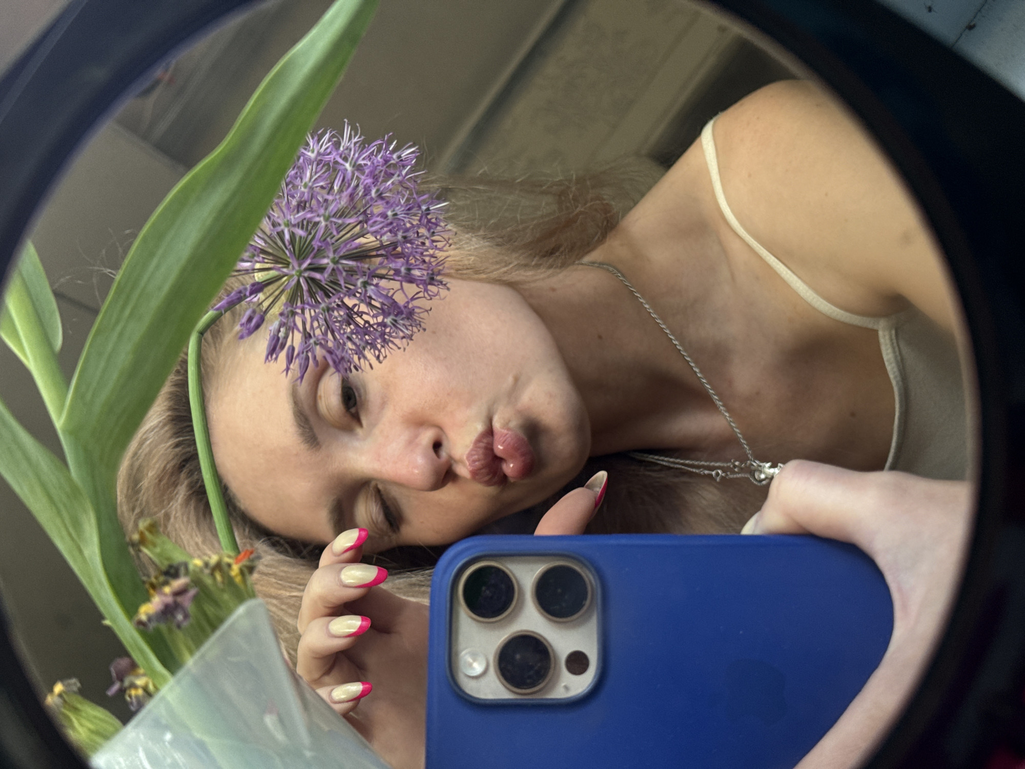 A young woman is taking a selfie in a mirror. She is holding a blue smartphone with three camera lenses. She has blonde hair, fair skin, and is wearing a white tank top and a silver necklace. She is pouting her lips. She is holding a flower in her hand with a purple flower head. The background is blurry but appears to be an interior space. There is no indication of the location being in the city of Chaikovsky, Russia.