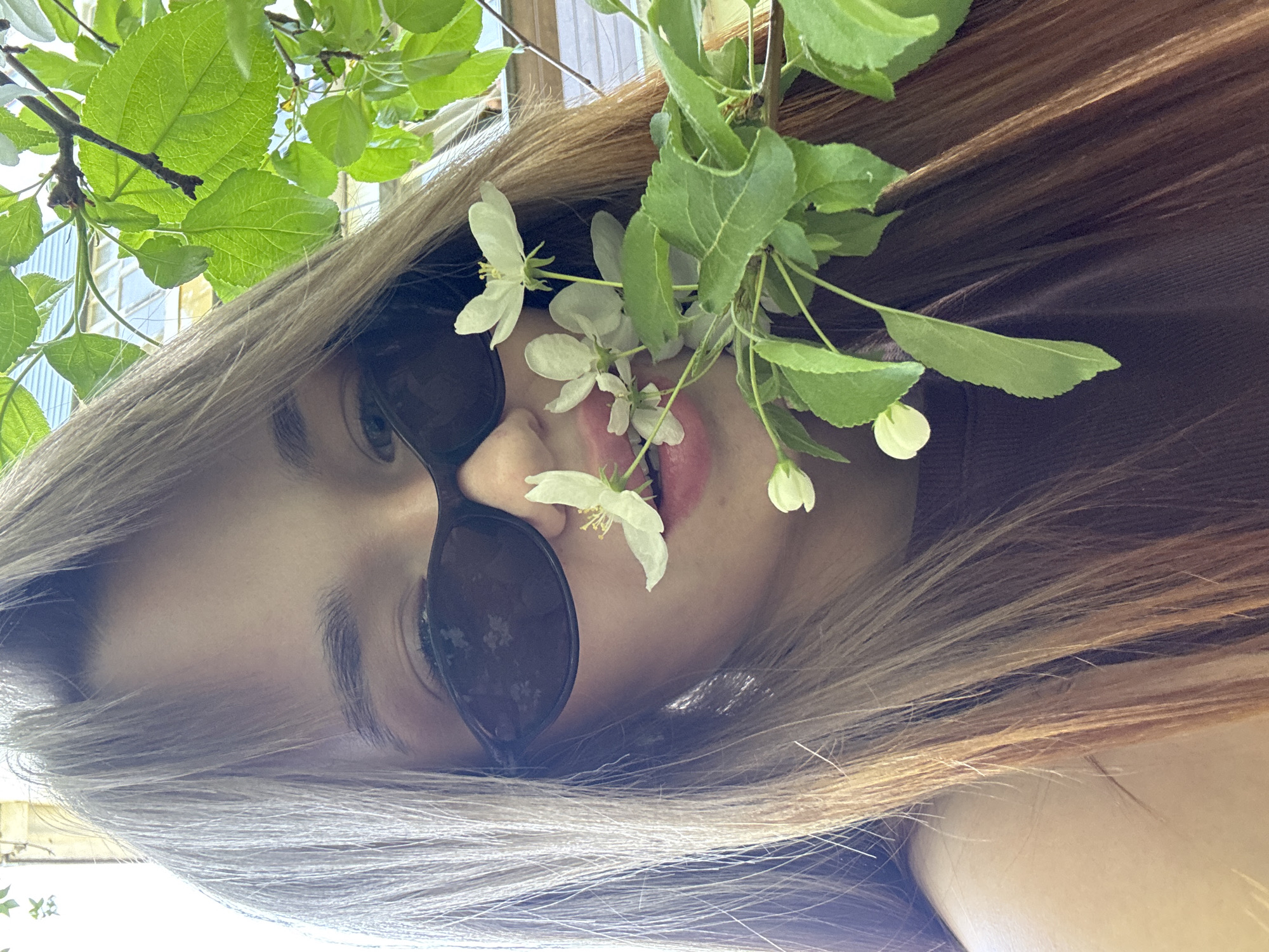 A young woman is photographed from a low angle, looking down at the camera. She is wearing dark sunglasses and has blonde and brown hair. A branch with green leaves and white flowers is held near her face. The background includes green leaves and a glimpse of a building. The image appears to have been taken outdoors in daylight. There is no specific information in the image to confirm the location as Chaikovsky, Russia.