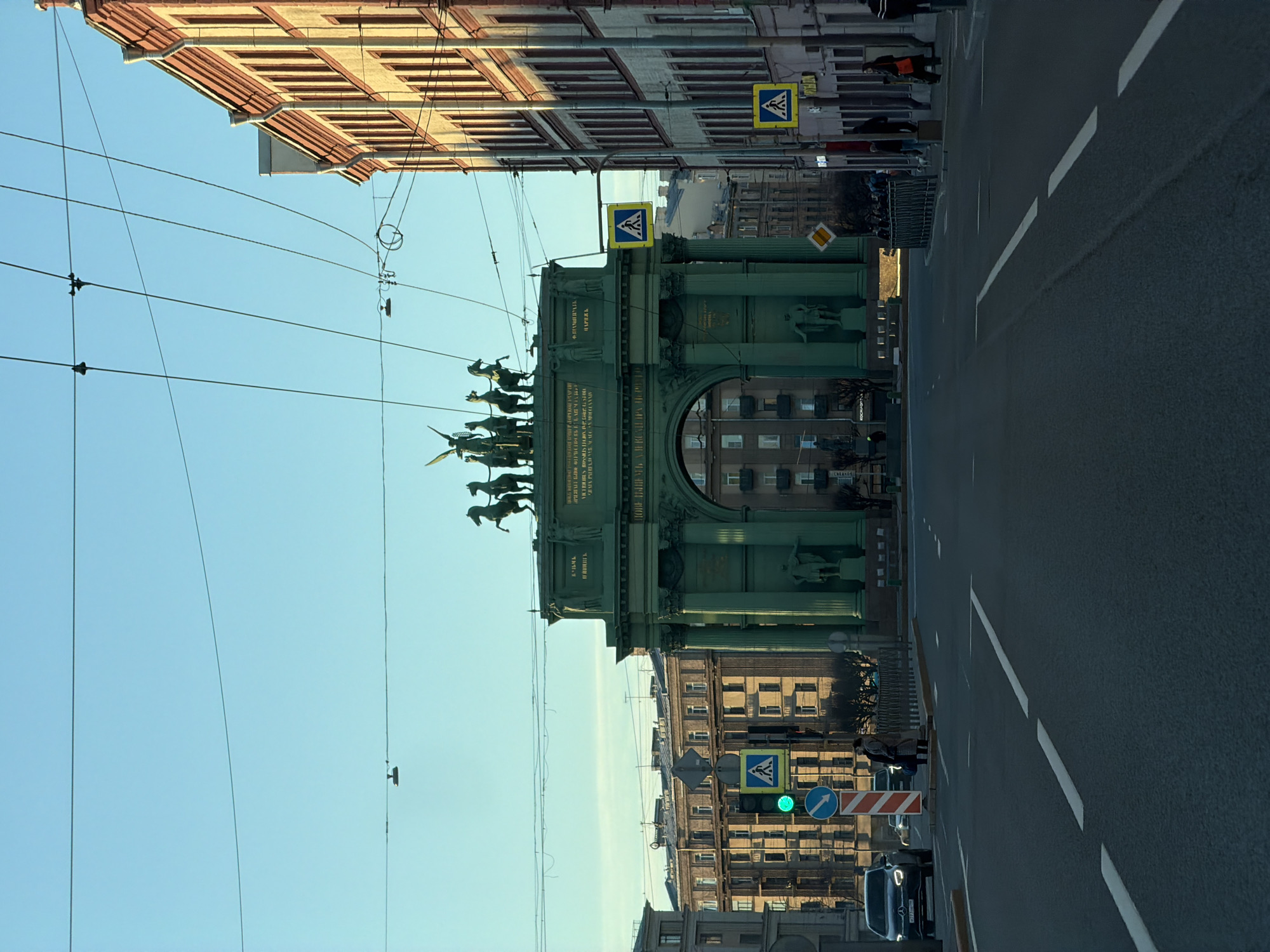 The image depicts the Narva Triumphal Arch in Saint Petersburg, Russia. It is a monumental arch of green color with sculptures and inscriptions, spanning across a wide road with white lane markings. On either side of the arch are buildings with yellow facades and rectangular windows. The sky is clear and blue. A pedestrian crossing sign is visible on both sides of the road. On one side, a traffic light shows green. Overhead power lines are strung across the view. A black vehicle is visible on the right side of the street.
