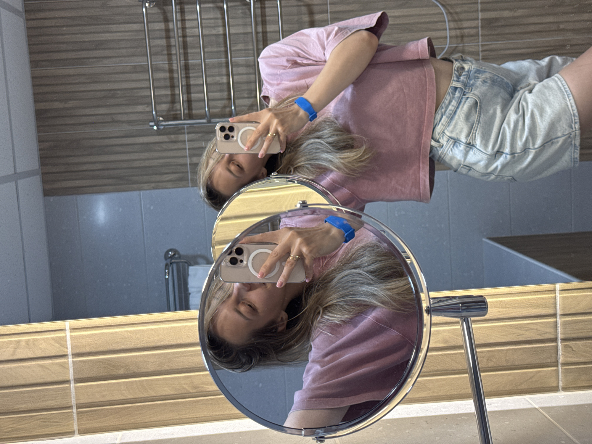 A young woman with blonde hair is taking a selfie in a mirror. She is wearing a pink cropped t-shirt and light-wash denim shorts. She has a blue wristband on her left wrist and a gold ring on her right index finger. The mirror is round and appears to be a makeup mirror on a stand. The background shows a tiled wall and a towel rack. The woman is leaning forward and smiling. The scene appears to be in a bathroom. The image does not contain any identifiable landmarks or features that would indicate the specific city of Nebug, Russia.