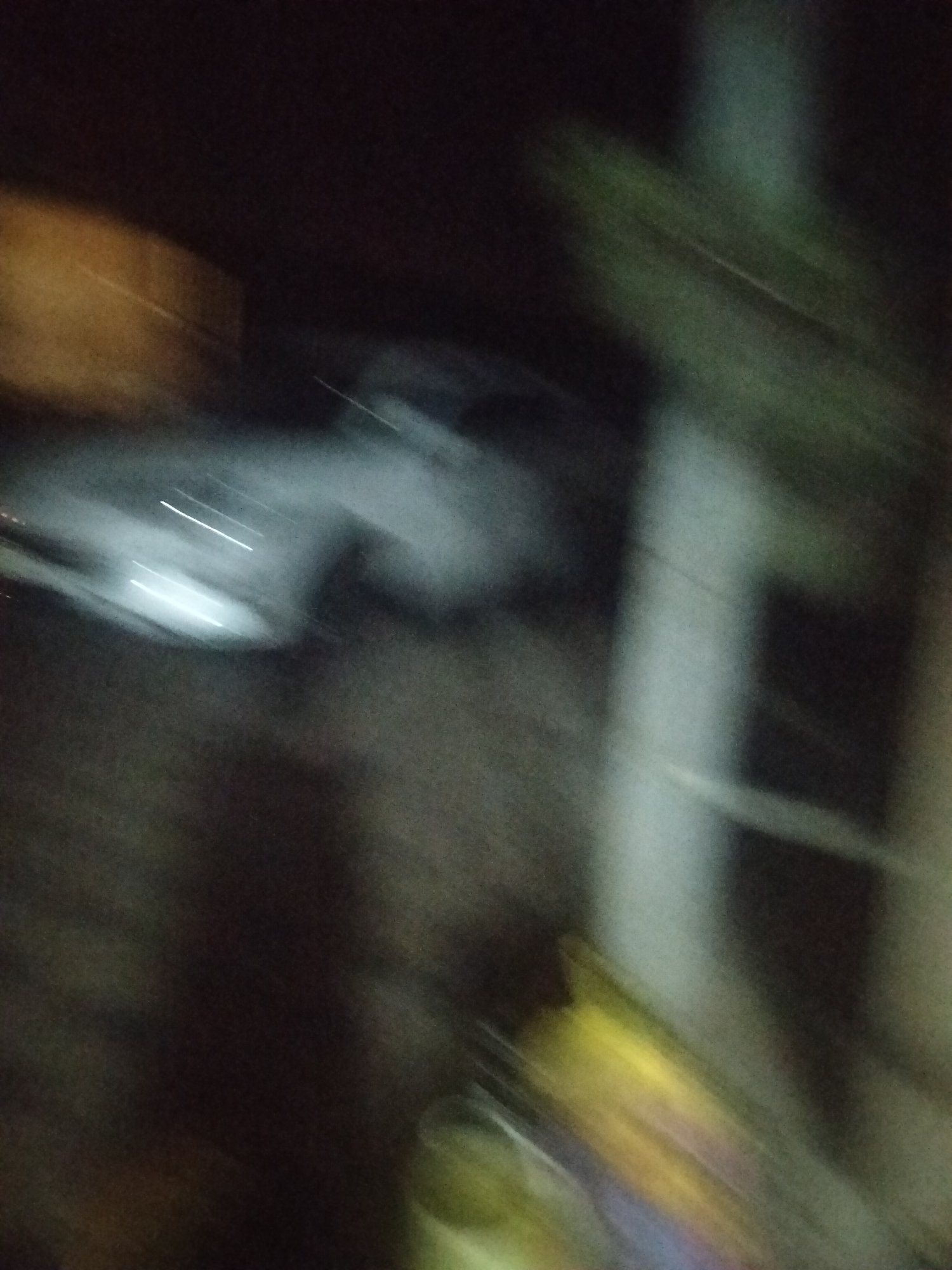 The image is a blurry, dark photograph that appears to be taken at night. The most prominent subject is a light-colored vehicle, possibly a car, with its headlights illuminated. The vehicle is in motion, indicated by light trails. The background is dark and indistinct, but suggests an outdoor environment. There are also some blurred areas of light and shadow that could be buildings or other urban elements. The overall impression is one of movement and low light conditions, characteristic of a street scene at night. The reference to Kagan, Uzbekistan, suggests this scene is located within that city.