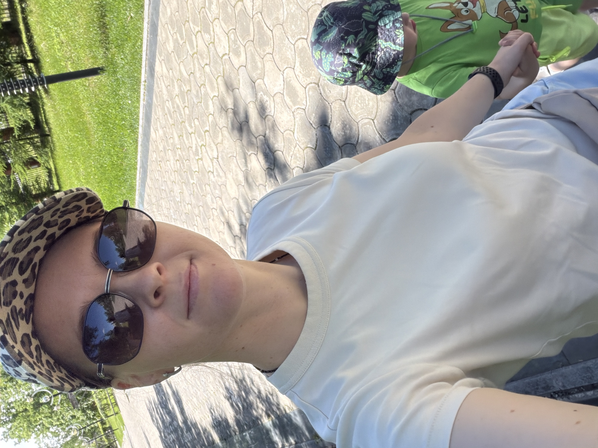 A woman wearing a leopard-print cap and sunglasses is taking a selfie in an outdoor setting. She has on a light-colored t-shirt. She is holding the hand of a young child who is wearing a green t-shirt with a cartoon dog print and a matching green patterned bucket hat. The child is also wearing green shorts. They are standing on a paved walkway made of interlocking stones. To the left of the walkway is a grassy area with trees and lampposts, suggesting a park or a well-maintained public space. The lighting indicates it is a sunny day. The background also shows a fence and more greenery, consistent with a park or an urban green space.
