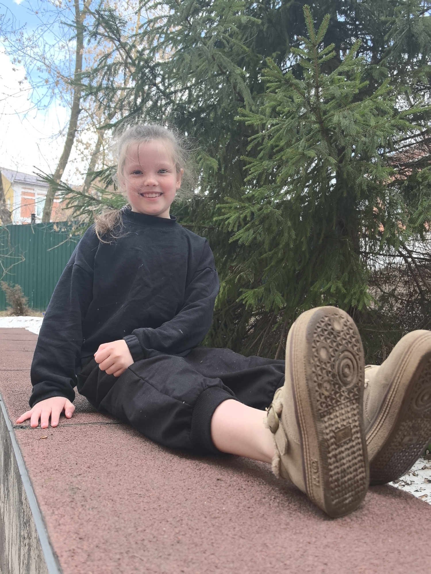 A young girl sits outdoors. She is wearing a black sweatshirt and pants, with dirty brown boots. She smiles at the camera. The location is in Nizhny Novgorod, Russia. The background includes a green fence, trees, and a building in the distance. Patches of snow are on the ground.