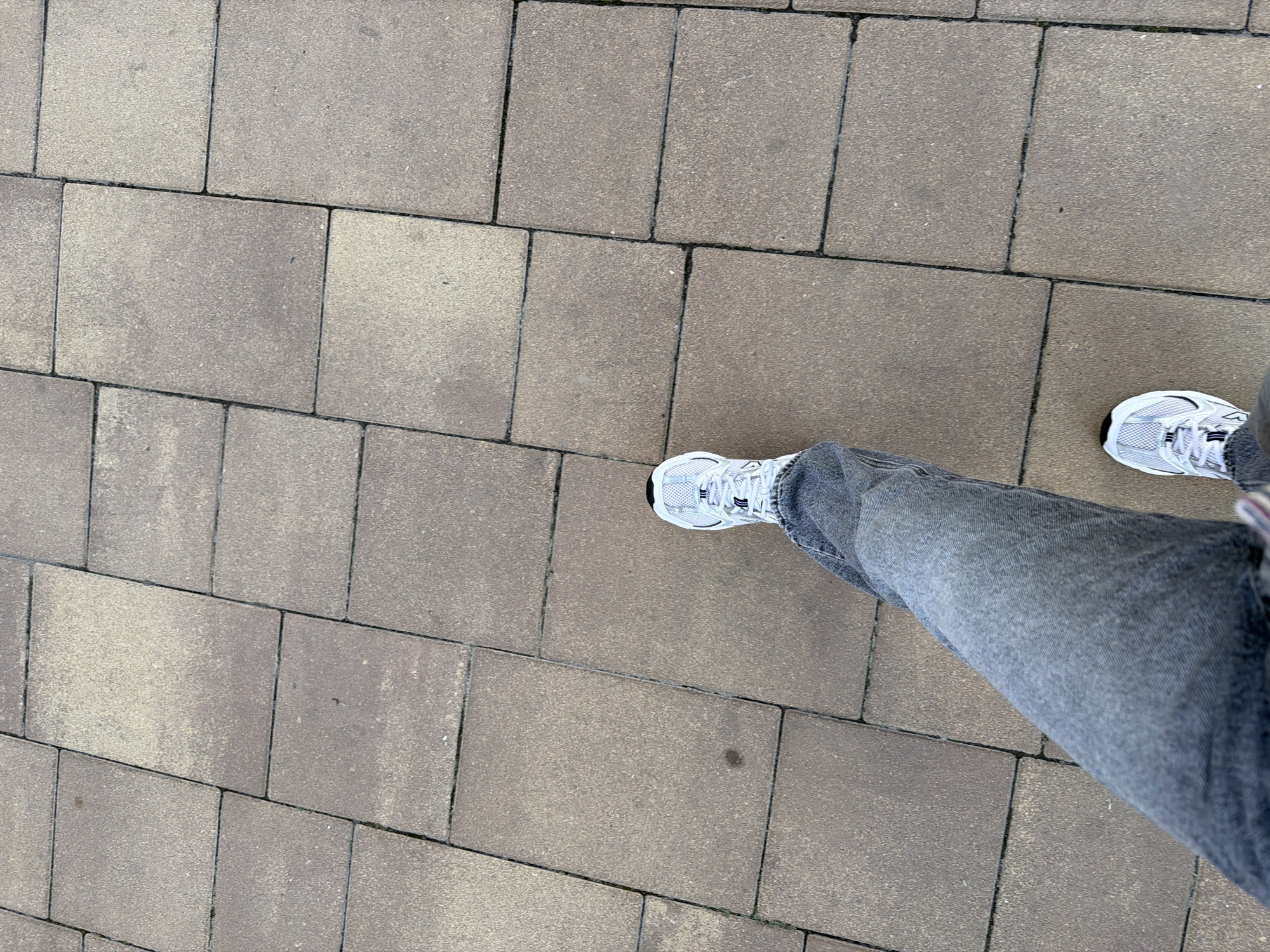The image shows a person's legs and feet from a first-person perspective, standing on a paved walkway made of rectangular brown and grey stone tiles. The person is wearing dark grey jeans and white athletic shoes with black and grey accents. The lighting suggests it is daytime, and the environment appears to be outdoors in an urban setting, consistent with a street or park. No specific landmarks or identifying features of Kyiv, Ukraine are visible in this image.