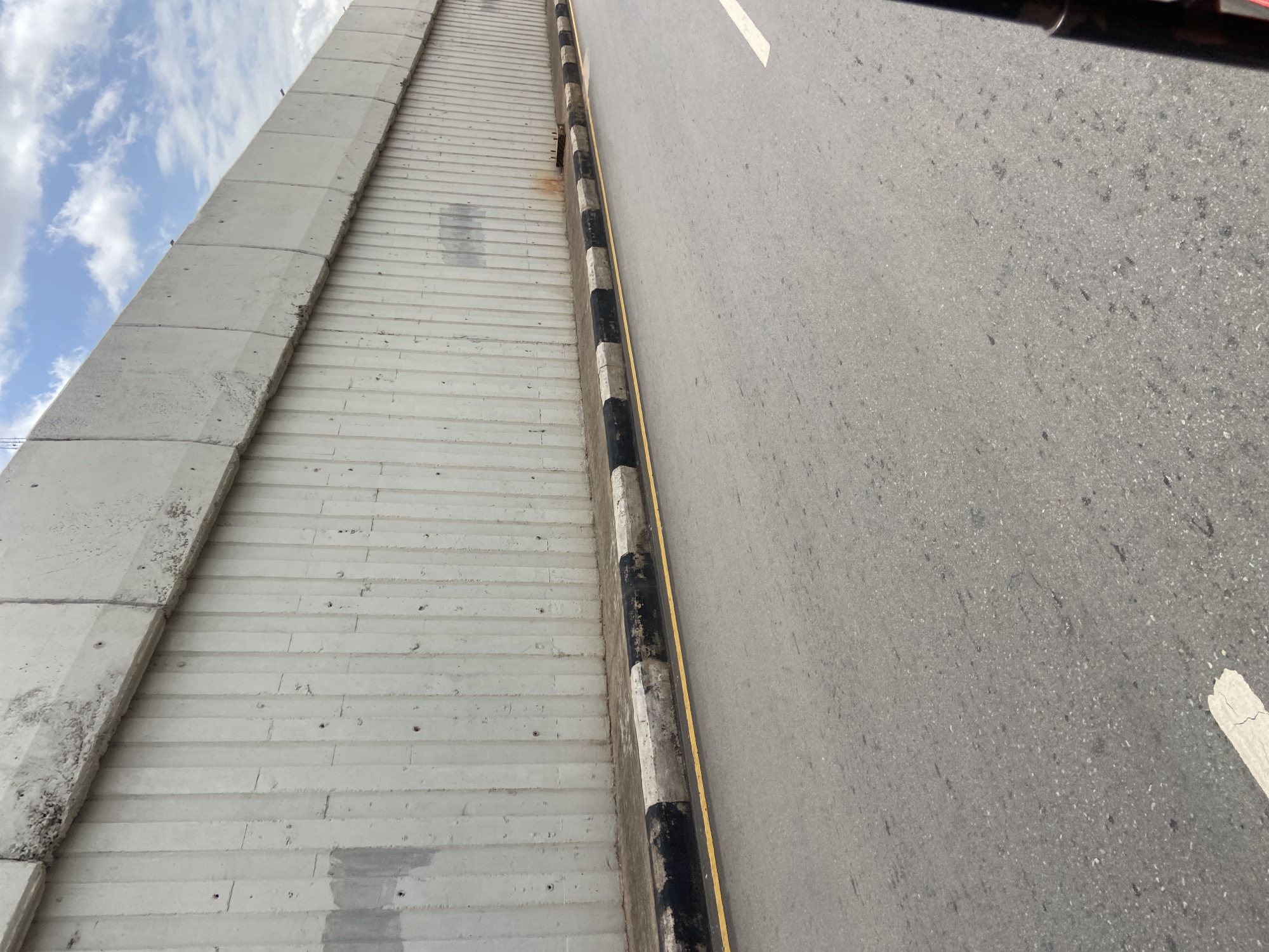 A low-angle, eye-level shot captures a section of a roadside in Jos, Nigeria, dominated by a tall, grey concrete barrier. This barrier, constructed from vertical concrete panels, forms a retaining wall, with visible signs of wear such as exposed aggregate and minor cracks. Its top edge is uneven, with some sections appearing chipped or eroded. Below the concrete barrier is a narrow strip of greyish ground, followed by a curb painted with alternating black and white stripes, suggesting a designated boundary. A thin yellow line runs along the edge of the asphalt road, separating it from the curb. The road itself is grey asphalt, showing some texture and scattered dark specks. A white line is partially visible on the road surface, indicating a lane marking. The sky above is partly cloudy, with a mix of white clouds and blue sky visible beyond the top of the concrete barrier. The overall scene depicts a common urban infrastructure element, likely part of a highway or elevated road system in Jos. No people or vehicles are prominently featured, focusing the image on the static elements of the environment.