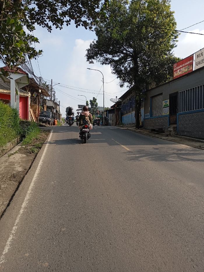 The image captures a daytime street scene in Pataruman, Indonesia, viewed from a slightly elevated, forward-facing perspective along an asphalt road. In the foreground-midground, a dark-colored motorcycle with a single rider is positioned centrally, moving away from the camera. The rider wears a light-colored head covering, a dark garment, and a backpack, with a brown helmet visible. The road features a solid white line delineating its left edge and partially visible yellow dashed lines further down the center.
On the left side of the road, a concrete curb separates the asphalt from a grassy verge. Beyond the verge, a series of buildings, some with red or pink architectural elements, are visible. A dark-colored utility vehicle is parked alongside the road in this area. Overhead, numerous utility poles support a dense network of power and communication lines spanning across the street.
On the right side of the road, commercial-style buildings are present. One building features a blue and grey facade with a local emblem, while another displays a red-bordered commercial sign with Indonesian text, including "WARUNG HARIMU" and "MASAKAN SUND." A large, mature tree with dense green foliage stands prominently on the right, casting shadows onto the road surface. Multiple streetlights are visible along the road. The sky is clear light blue with scattered white clouds, indicating bright daylight conditions. Further down the road, additional motorcycles are visible, also traveling in the same direction.