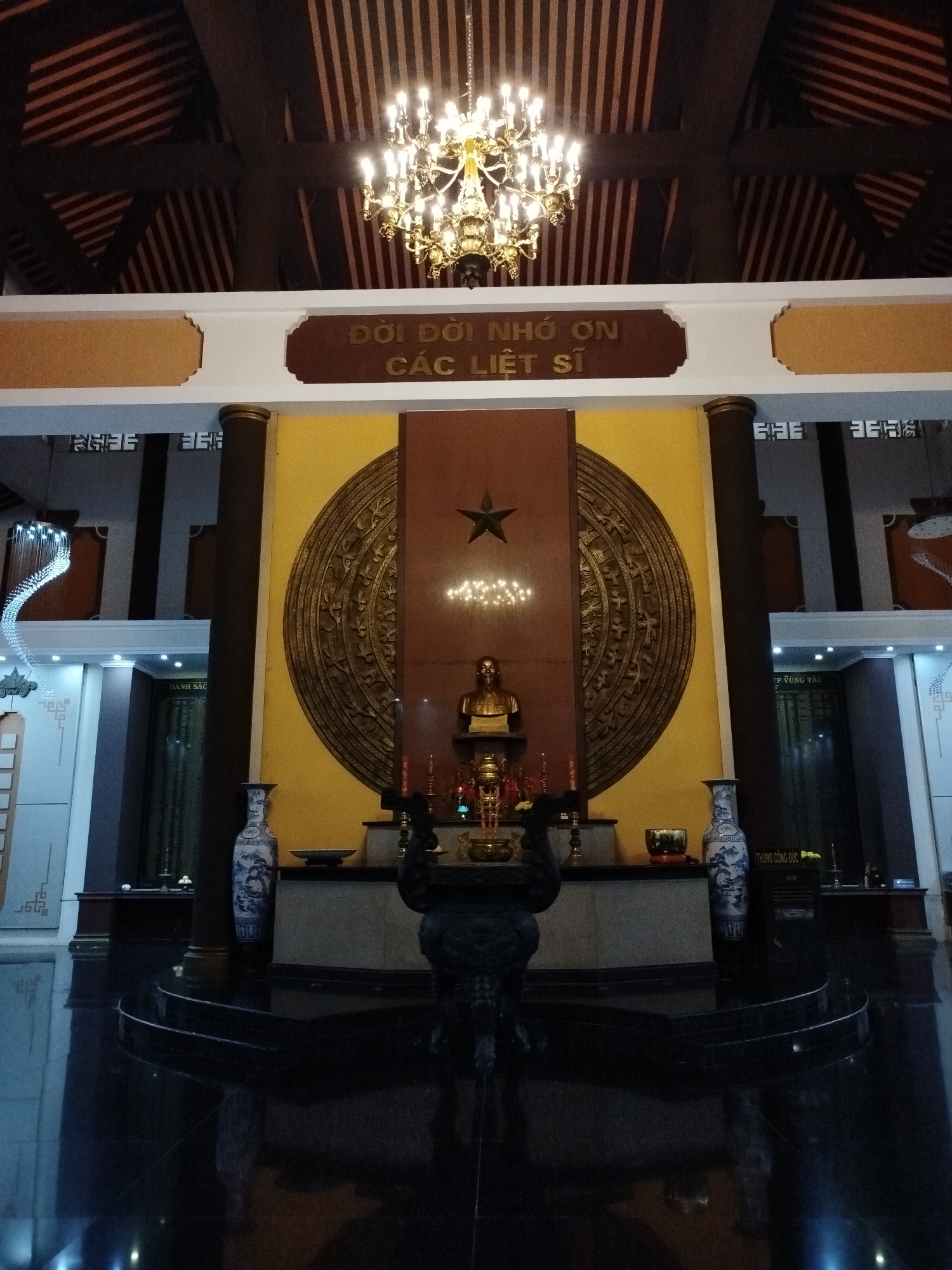 This is an indoor scene featuring a memorial in what seems to be a temple or a hall of remembrance. The scene is centered around a bronze gong, ornately decorated, hung on a yellow wall, overlaid with a brown rectangular backdrop bearing a black star at the top. In front of the gong, there's a bust of a person, possibly a historical figure, placed on a pedestal. Beneath the bust, there's an altar with candles, incense burners, and other religious items.
On either side of the altar are tall, decorative vases in light blue and white. The scene is flanked by dark, cylindrical pillars. Above the gong, a sign with brown letters on a light brown background reads "ĐỜI ĐỜI NHỚ ƠN CÁC LIỆT SĨ".
The floor is highly polished and reflective. At the top, a large, ornate chandelier hangs from the ceiling. The ceiling appears to be constructed of wooden beams, and the upper walls are decorated in white, gray, and beige colors. In the background, there appear to be columns or informational displays. Based on the text visible in the background and the memorial nature of the scene, this is likely a memorial hall for those who lost their lives. Without more details, it is hard to ascertain the exact location within Ho Chi Minh City.