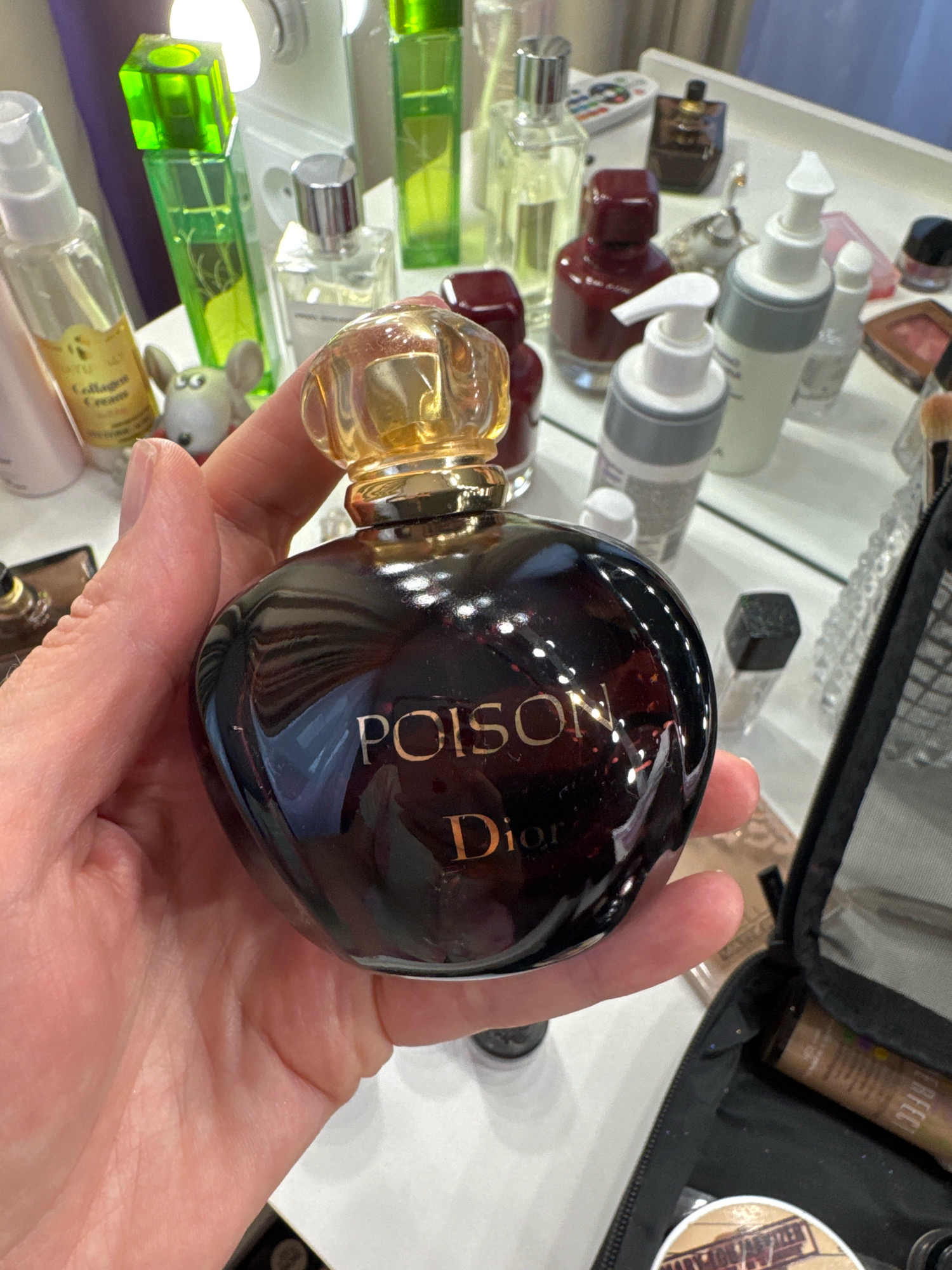 A hand holds a dark glass perfume bottle with a gold cap. The bottle has "POISON" and "Dior" printed on it in gold lettering. In the background, on a white surface, are several other cosmetic items, including bottles of perfume, lotion dispensers, and a makeup bag. The items are arranged on what appears to be a vanity or dressing table. The scene is well-lit, possibly by natural light or artificial lighting from above. There is no indication of the city of Sinyavino, Russia.
