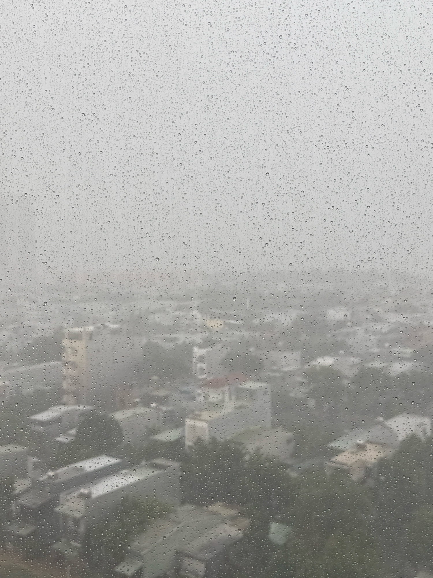The image is an exterior, medium shot of a cityscape viewed through a window covered in raindrops. The foreground is dominated by numerous water droplets of various sizes obscuring the view of the city in the background, which appears hazy and muted due to the rain and the glass. Buildings, primarily residential with pitched roofs, are visible through the water-streaked glass. Some structures have lighter-colored roofs, while others are a darker grey or blend into the overcast environment. Green foliage, likely trees, is interspersed between the buildings, suggesting a suburban or mixed urban setting. The overall impression is one of a rainy day in an unidentified urban area, with the water on the window creating a diffused and atmospheric perspective.