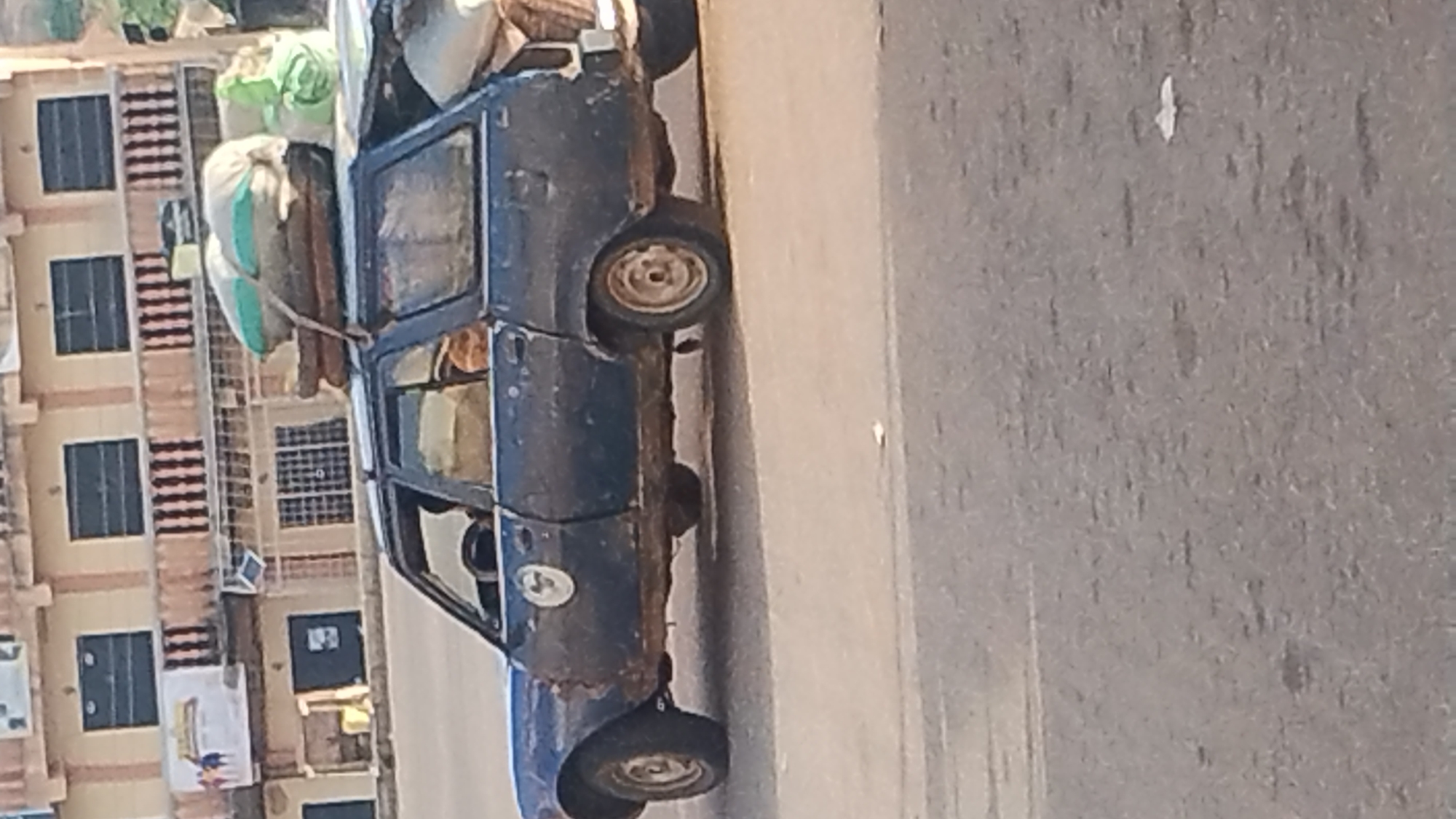 A dilapidated blue car is heavily laden with bags on its roof. The car is parked on a grey paved surface resembling a road. A multi-story building with multiple windows is visible in the background, suggesting an urban location. The scene seems to be set in the vicinity of New Stadium, Nigeria.