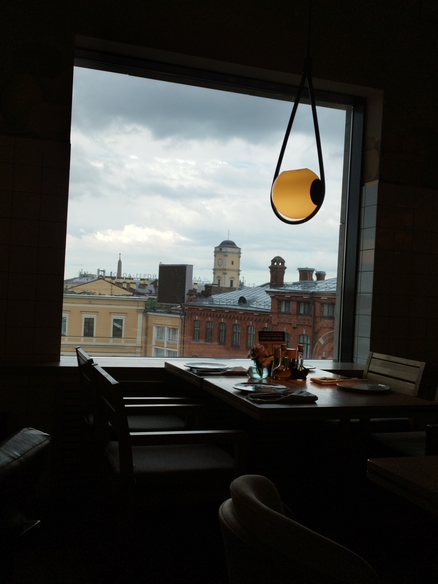 The image shows a view from inside a restaurant in Saint Petersburg, Russia, looking out a large window at the city skyline. The window frames a cloudy sky and several buildings, including one with a clock tower. Inside the restaurant, a table is set for a meal with plates, silverware, and a vase of flowers. A modern pendant light with a yellow bulb hangs from the ceiling. The lighting in the restaurant is dim, creating a silhouette effect of the furniture. The overall atmosphere is one of a cozy and dimly lit dining space with an urban view.