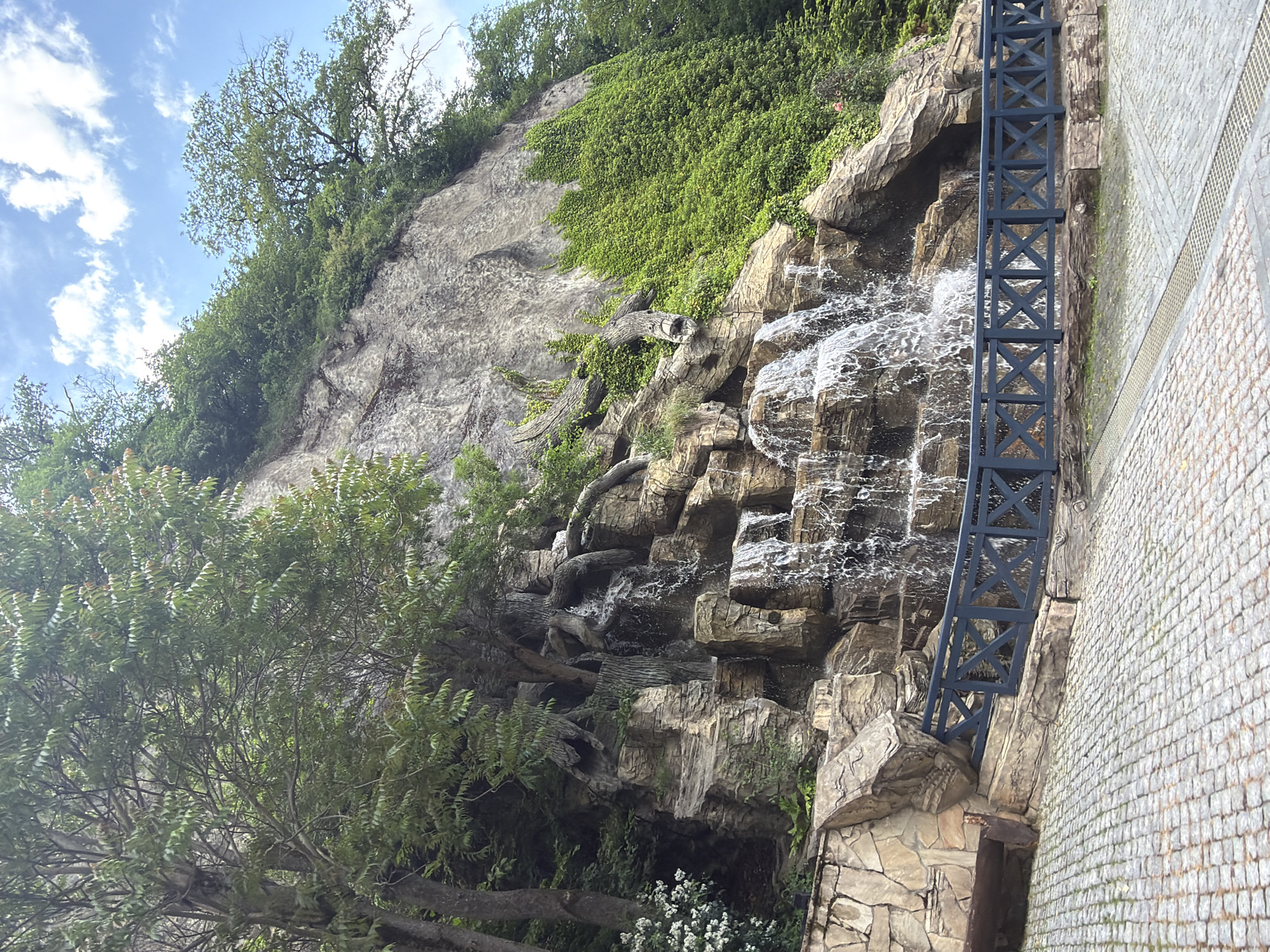 The image shows a multi-tiered waterfall cascading down a rocky cliff face. The cliff is covered in lush green vegetation, including ivy. Trees with exposed roots are growing from the top of the cliff and overhang the waterfall. A dark blue wooden railing with an 'X' pattern runs in front of the waterfall, separating it from a cobblestone paved area. A wooden bench is visible to the left of the railing. The sky above is blue with scattered white clouds. This scene is located in Borjomi, Georgia.