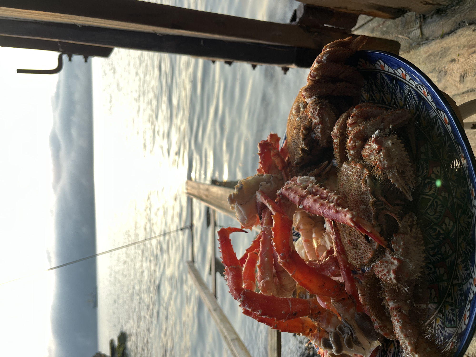 In Sovetskaya Gavan', Russia, at sunrise, a plate of cooked crab legs and whole crabs is presented on a wooden surface. The crabs, vibrant red and spiky, are arranged on a blue and white patterned plate. In the background, the calm sea reflects the warm glow of the rising sun, with a distant ship and a misty shoreline visible. A wooden structure and a fishing rod are positioned to the right and center of the frame respectively, adding to the outdoor, seaside ambiance. The scene suggests a fresh catch enjoyed at the water's edge.