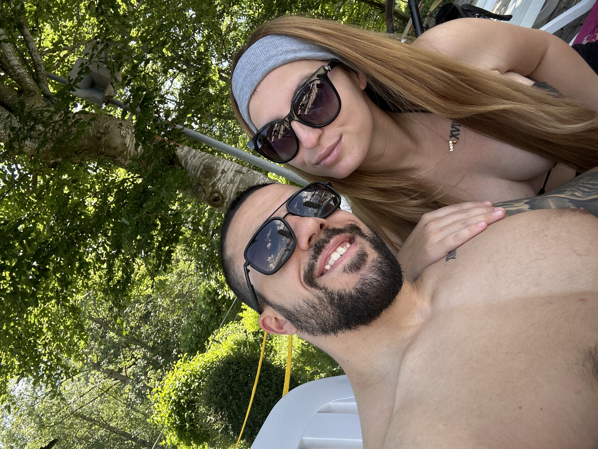 A shirtless man with a beard and sunglasses smiles at the camera, while a woman wearing sunglasses and a headband is next to him, with her arm resting on his shoulder. They are sitting outdoors in a wooded area, with trees and foliage surrounding them. The man has a tattoo on his chest. The woman is wearing a necklace and has a tattoo on her clavicle. The scene appears to be a relaxed, outdoor setting. There is a white plastic chair visible in the foreground.
