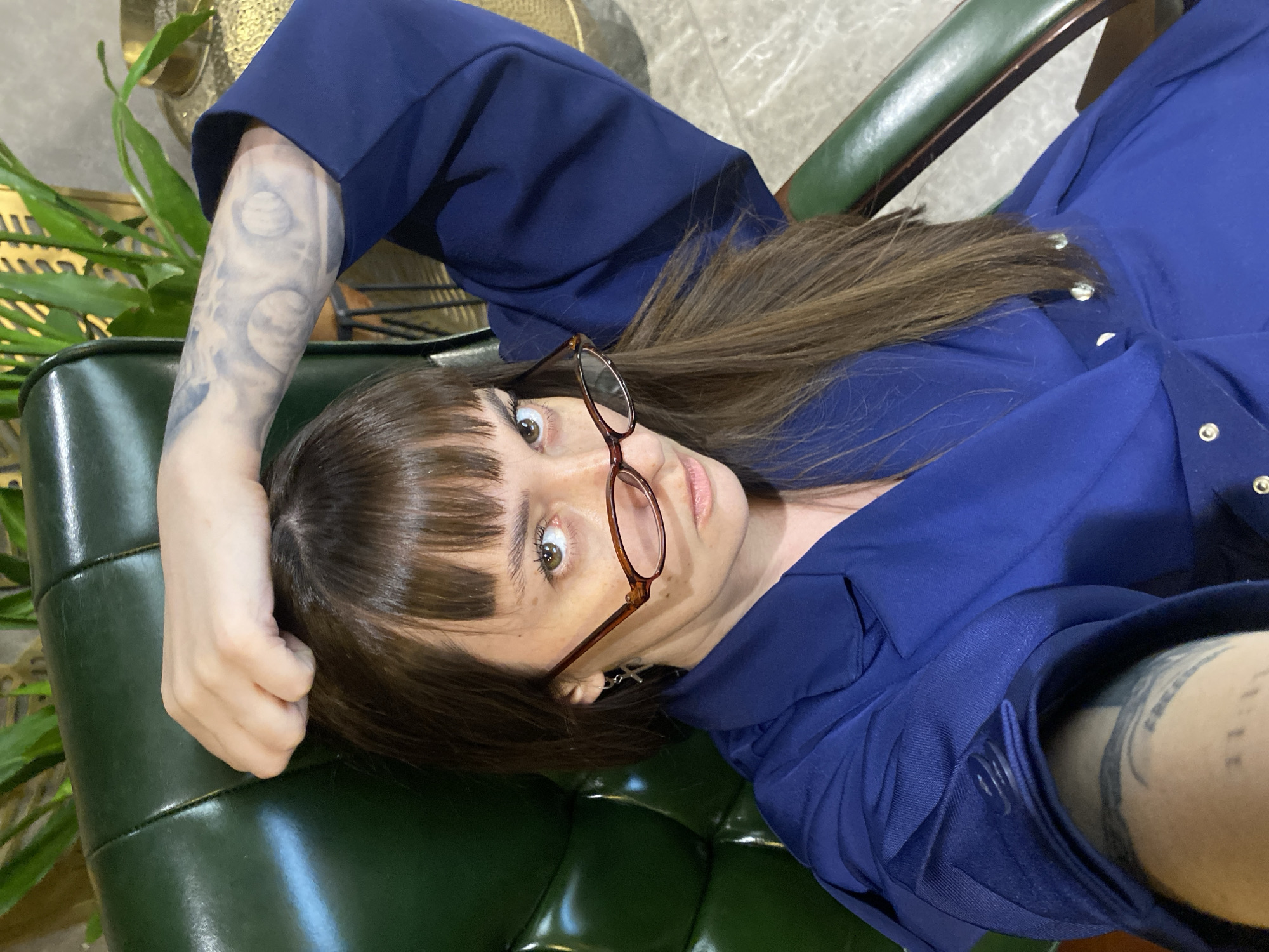 A person with brown hair, bangs, and eyeglasses is lying on a green padded chair. They are wearing a dark blue, button-down shirt or dress. Tattoos are visible on both arms. The environment includes an indoor space with a plant in the background. There is no information available that would allow me to infer or determine if this image was taken in Votkinsk, Russia.