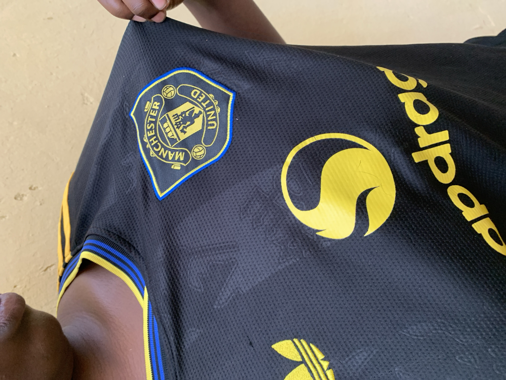 A black soccer jersey with yellow accents is held up by a person in Rafin Par, Nigeria. The jersey features the Manchester United logo on the left chest and the Adidas logo at the bottom. A logo resembling a swirling letter "S" and the inscription "adrag" are printed on the right side of the chest. The sleeve cuffs have yellow and blue stripes. The jersey appears to be made of a textured material. A portion of a person's face and arm are visible. The background is a plain, light-colored wall.