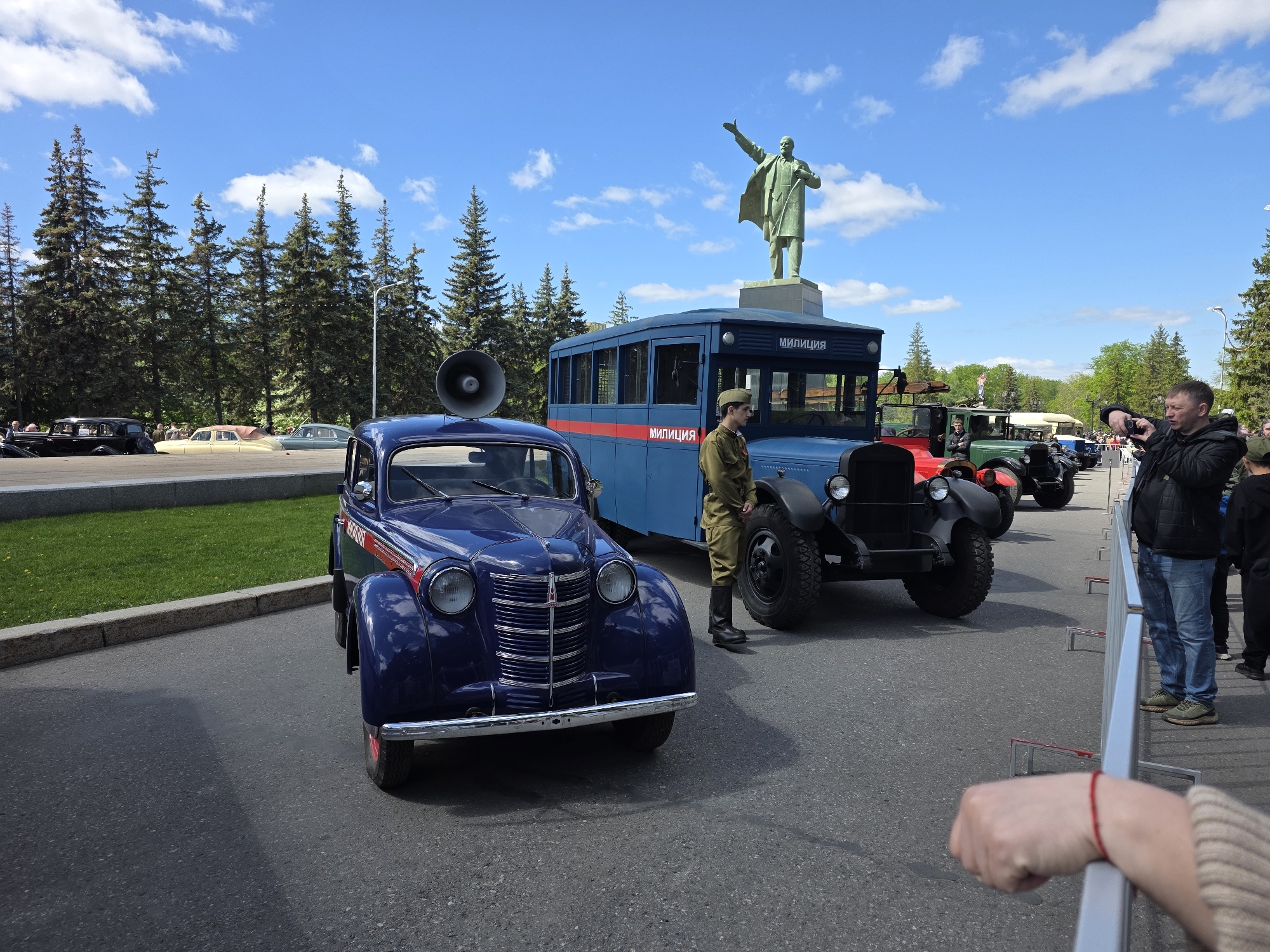 The image shows a collection of vintage vehicles on display in Ufa, Russia. In the foreground is a dark blue Moskvich 400 car with a large megaphone on its roof. Behind it stands a blue truck-bus with the word "милиция" (police) written on its side. A person in an old military uniform stands next to the truck. In the background, a statue of Lenin is visible atop a building. A man to the right is taking a picture with his phone. The scene takes place on an asphalt road, with trees and a blue sky with scattered clouds above.
