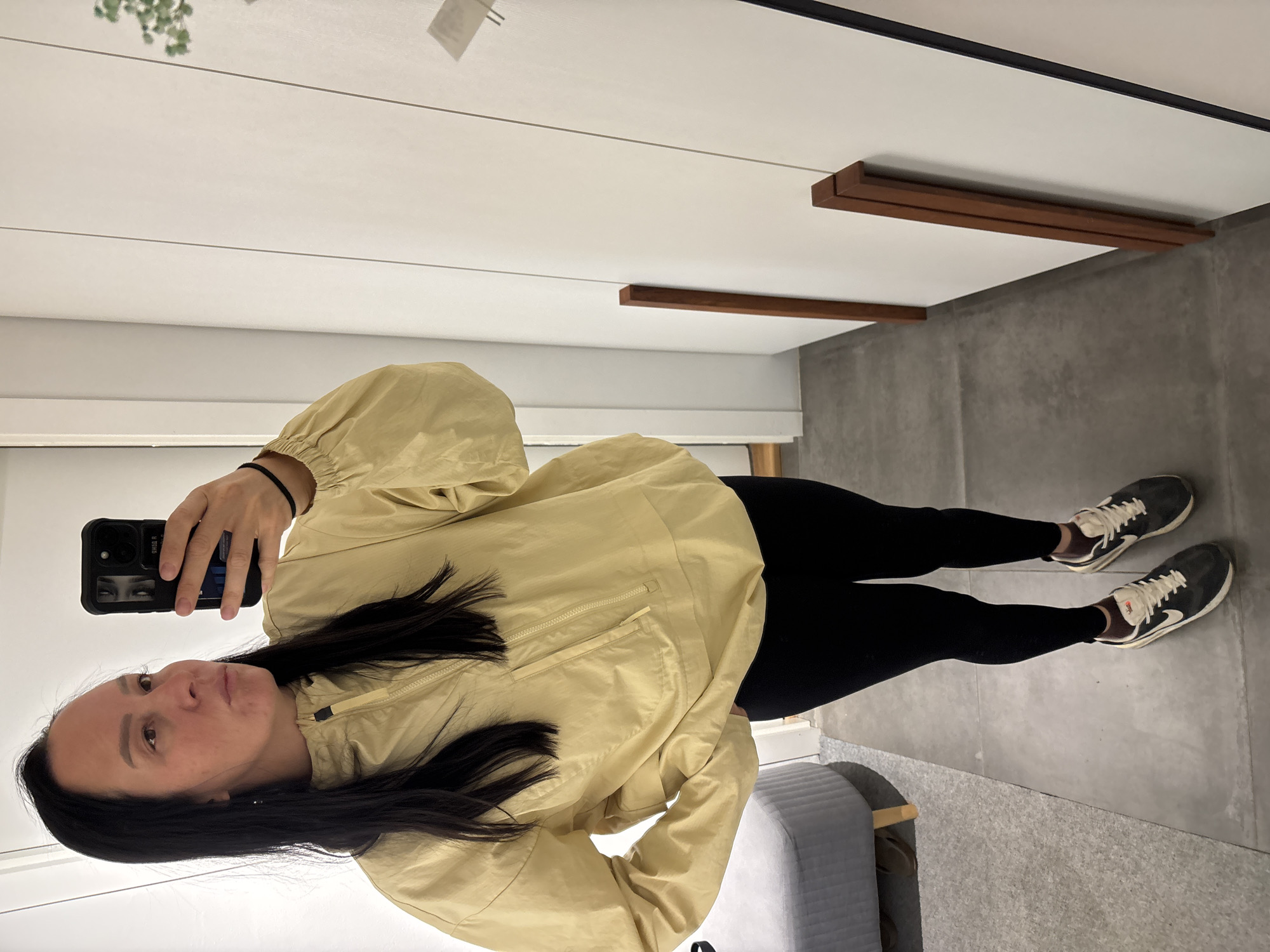 A woman is seen taking a selfie in what appears to be an indoor setting, possibly a dressing room or shop. She is wearing a light yellow jacket, black leggings, and dark sneakers with white laces. She is holding a smartphone up, the screen visible showing a reflected image of her face. The background includes a white paneled wall with dark trim at the top and bottom. A wooden decorative beam is visible in the upper right corner. The floor is a neutral gray color. A small, light gray stool or chair is partially visible in the lower right corner. There is no verifiable evidence to place this scene specifically in the city of Chaikovsky, Russia based on the image alone.