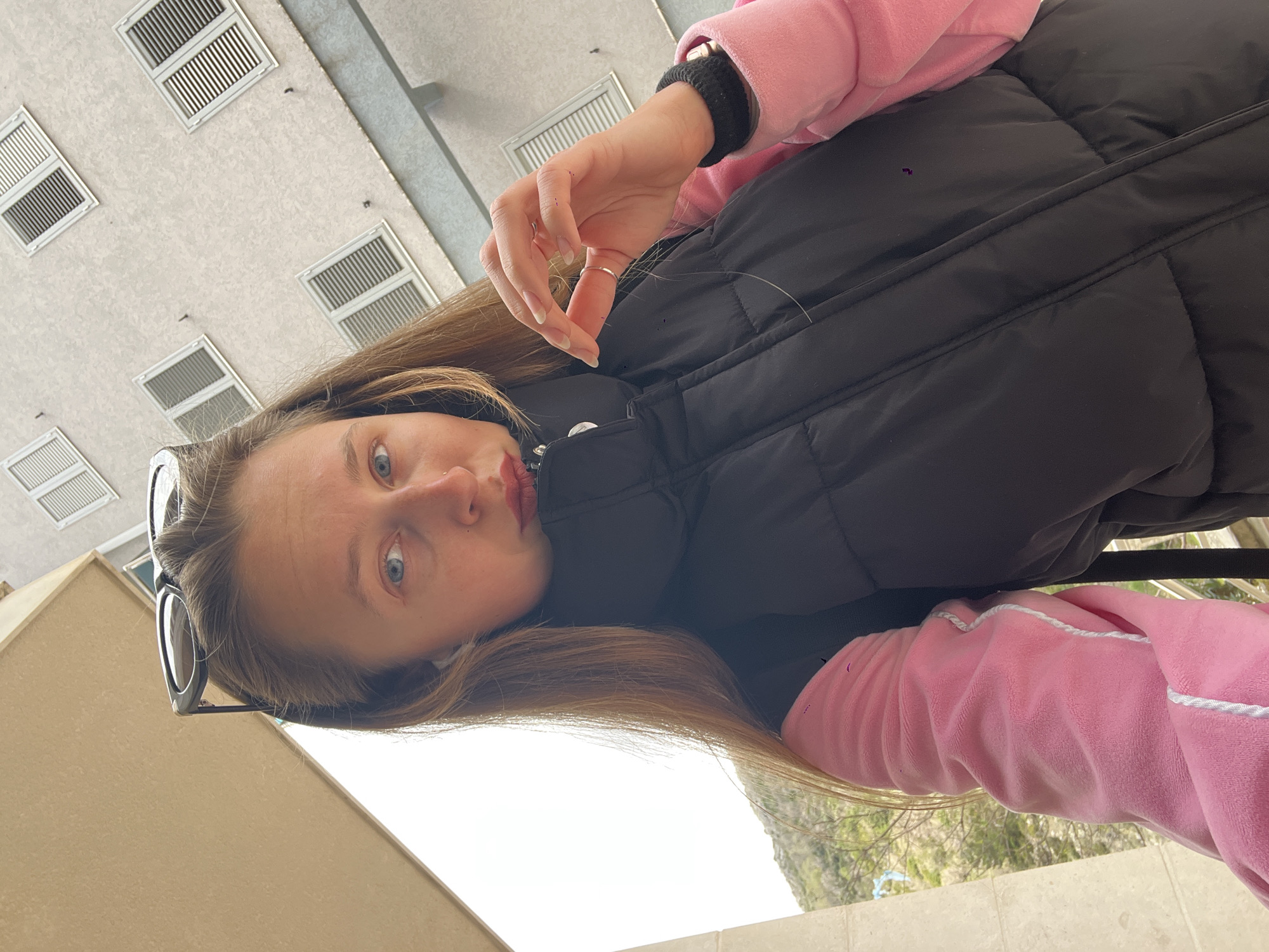 A woman with long blonde hair, blue eyes, and a nose ring is looking at the camera. She is wearing a black vest over a pink jacket with white trim. Her left hand, adorned with a silver ring, is raised. Sunglasses are tucked into her hair. The background includes a beige wall with windows, foliage, and the sky. The location is Rafailovići, Montenegro.