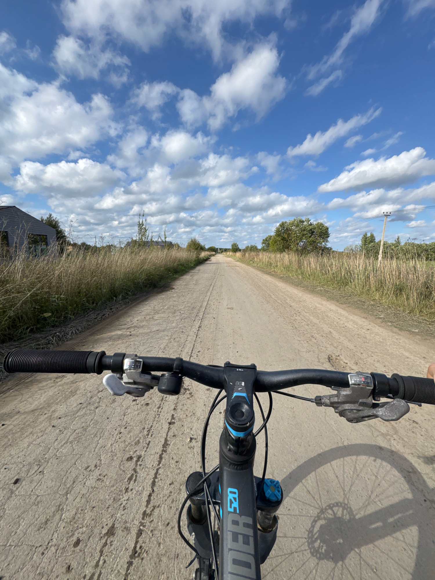 A first-person perspective shot captures a cyclist's view of a dirt road stretching into the distance. The road is lined with dry grass and some foliage on both sides, with a building visible on the left and trees and a utility pole further ahead. The sky above is a vibrant blue, dotted with fluffy white clouds. The cyclist's hands are on the handlebars of a black bicycle, and part of the front wheel and fork are visible. The scene is set in Kempelevo, Russia, under a bright, sunny sky.