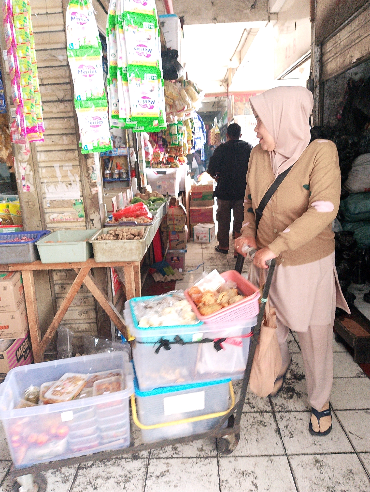 This is a forensic, matter-of-fact description of an outdoor market scene in Lembang, Indonesia. The foreground features a woman wearing a light brown hijab and a matching long-sleeved cardigan, as well as light beige pants and dark blue flip-flops. She is pushing a small trolley laden with transparent plastic containers. The containers hold various food items, including what appears to be fried snacks in a pink basket on top, and other packaged goods. The trolley has a black handle and a brown bag hanging from it.
Behind the woman, a man in a black shirt and brown pants is visible, walking away from the camera. The background depicts a bustling market setting. Stalls are lined with shelves displaying various products. Hanging from a stall on the left are several colorful packages, possibly snacks or small goods. In the center, a stall features shelves with food items, including red chilies and green vegetables in plastic containers. There are also wooden tables with more produce and packaged goods. The ground is paved with textured tiles, some of which appear wet. Boxes and various market wares are stacked around the stalls. The overall environment suggests a typical Indonesian market, characterized by its vibrant and crowded atmosphere.