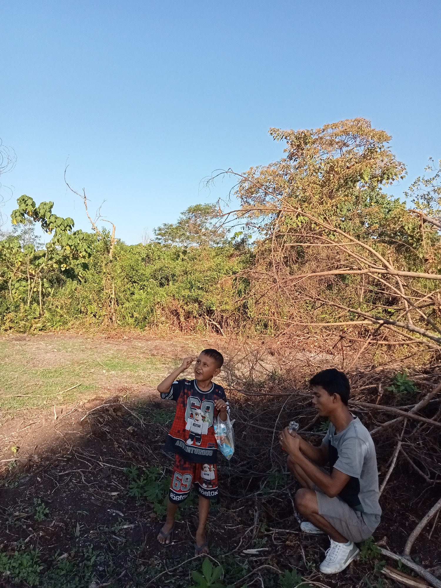 In Kualu, Indonesia, under a clear blue sky, a young boy wearing a colorful t-shirt and shorts with "Roblox" motifs stands to the left, holding a plastic bag. To his right, a young man crouches, holding a bottle in his hands. They are surrounded by dry branches and lush green foliage. The ground is covered with dry leaves and sparse grass.