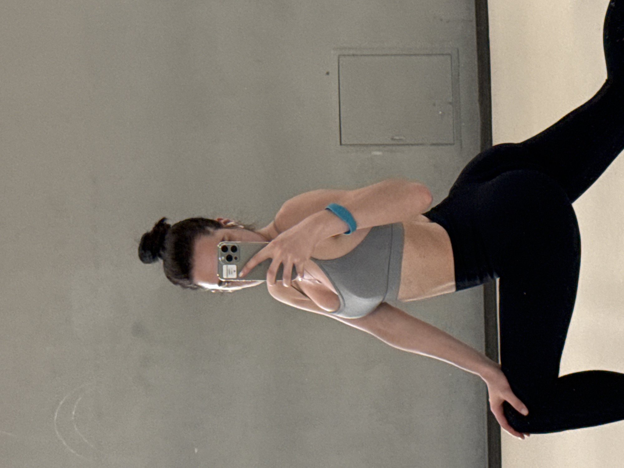 A woman is taking a mirror selfie. She is wearing a gray sports bra and black leggings. She has a bun in her hair and is holding her phone in her left hand. There is a blue bracelet on her left wrist. The background is a gray wall with a rectangular access panel. The location appears to be a room with a mirror. The city and state are unknown.