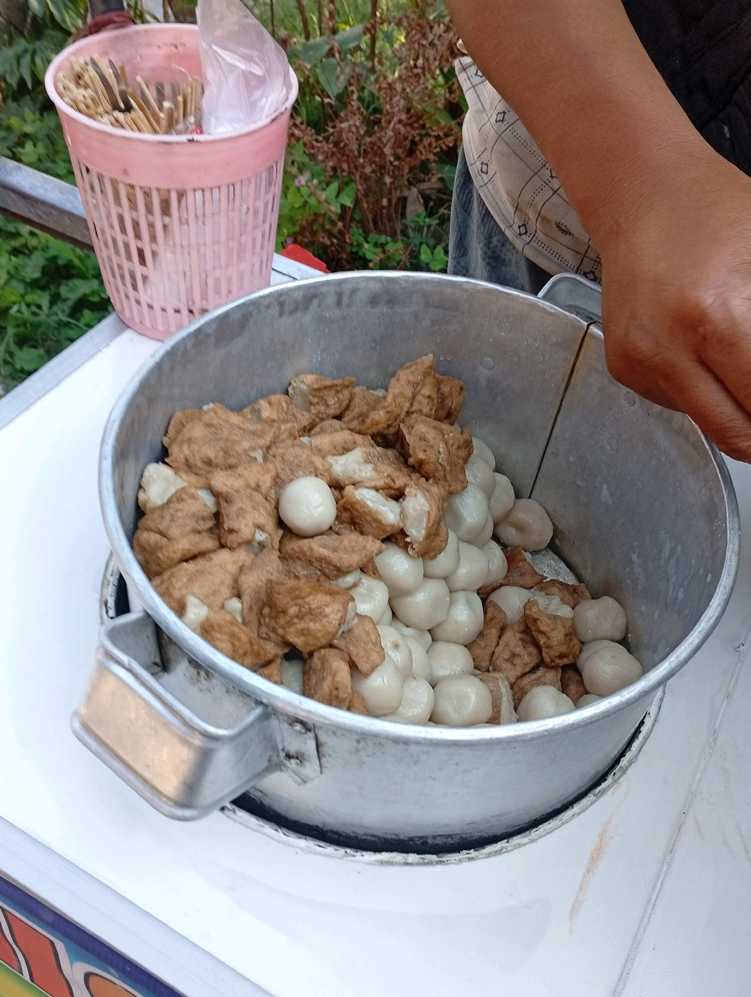 This is a close-up, medium-angle shot of a street food vendor's cooking setup. The primary focus is a large, dented aluminum pot filled with food. The food consists of two main types: small, pale, spherical items and irregularly shaped, browned chunks that appear to be fried tofu or some other fried savory item. The items are mixed together. A person's forearm and hand, with tanned skin, are visible on the right side of the frame, reaching towards the pot. To the left of the pot is a pink, plastic basket containing a bundle of wooden skewers, some of which are covered by a translucent plastic bag. The background shows out-of-focus green foliage and dry brown vegetation, suggesting an outdoor setting. The pot is placed on a white surface, possibly a cart or table, which has a colorful, patterned edge at the bottom of the frame. The scene is likely located on a street or sidewalk in Salatiga, Indonesia, a common setting for such food vendors.