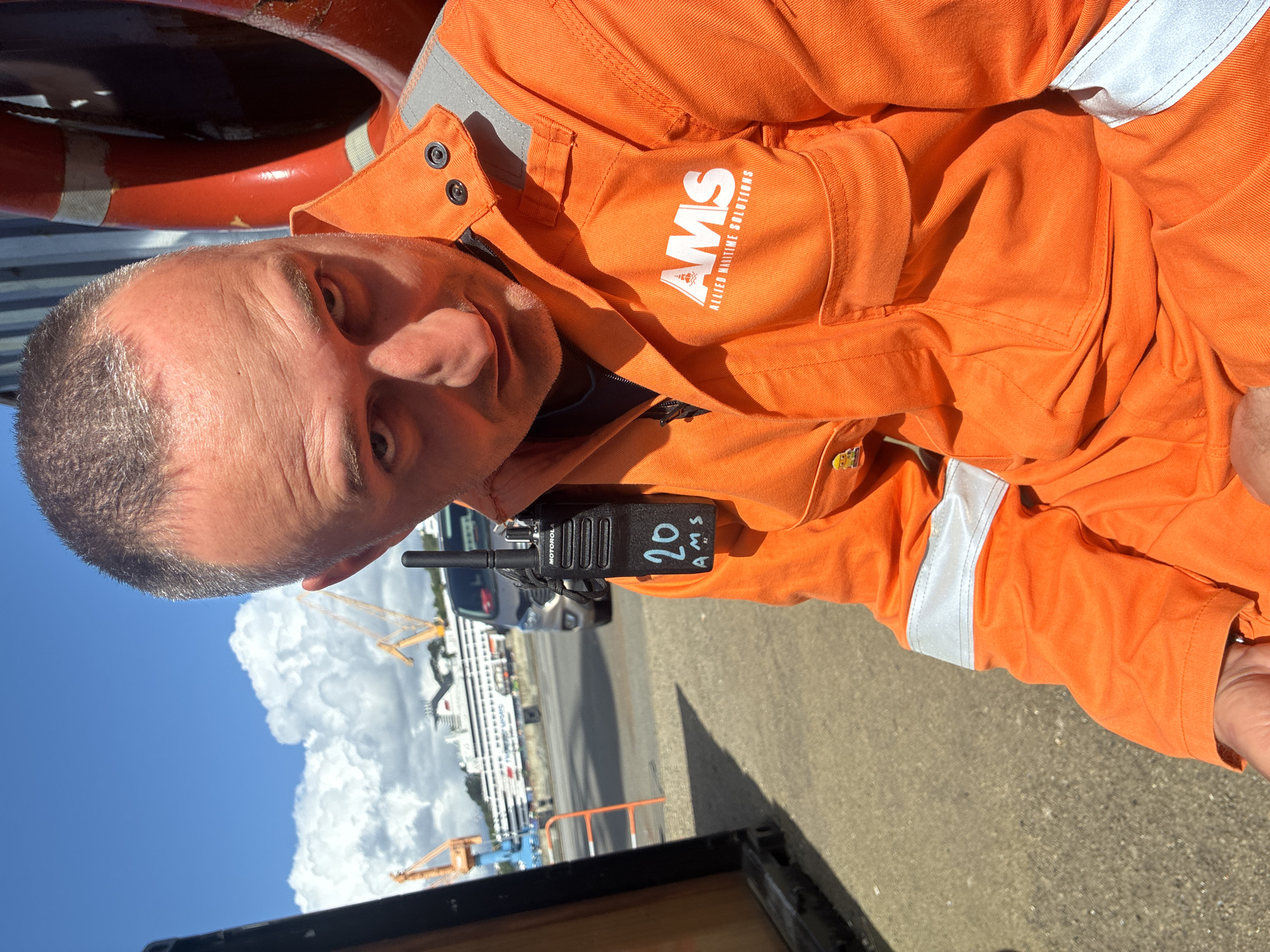 A man wearing an orange jumpsuit with "AMS Allied Maritime Solutions" printed on the upper right side is shown in a selfie. He is looking at the camera. He is wearing a black Motorola radio with "20 AMS" written on it. The backdrop shows a sunny day with some large clouds in the sky. In the background, a body of water is visible, along with fencing, vehicles, and cranes. These features suggest the location is the harbor in Brest, France.