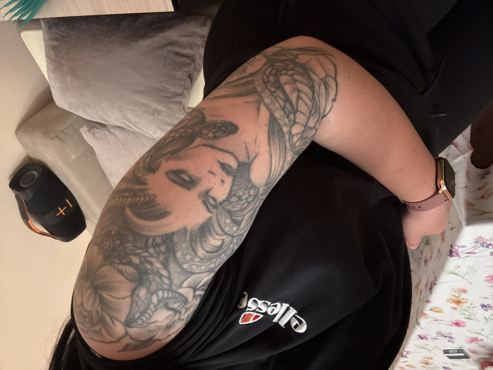 The image shows a person seated, wearing black sweatpants with the "Ellesse" logo visible. The person's left arm is heavily tattooed, featuring a detailed design with a woman's face, floral elements, and a dragon or snake. They are wearing a pink smartwatch on their right wrist. A speaker is visible to the left of the frame. There are pillows and what appears to be a couch in the background. The person is seated at a table with a floral patterned tablecloth. The image was likely taken indoors. There is no information to suggest the location is Plovdiv, Bulgaria.