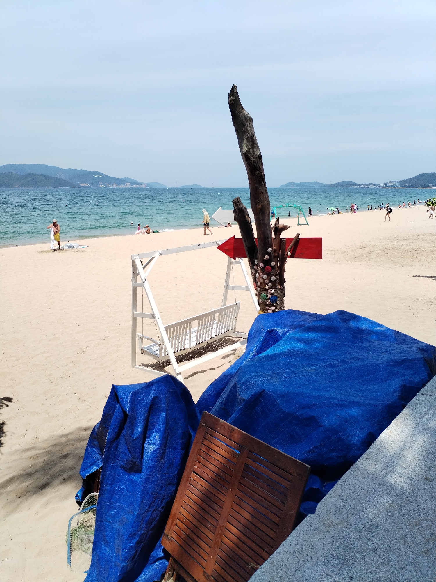 This is a photograph of a beach in Nha Trang Ward, Vietnam. In the foreground, there are several large blue tarps covering objects, along with a wooden slatted object. A white swing set is positioned on the sand behind the tarps. To the left of the swing set, a rustic wooden post adorned with shells stands prominently. A red arrow sign is affixed to this post, pointing to the left. The beach is populated with people, some of whom are walking or sitting near the water's edge. The sea is a clear blue, and in the distance, hills and buildings are visible along the coastline. The sky is overcast with a few scattered clouds.