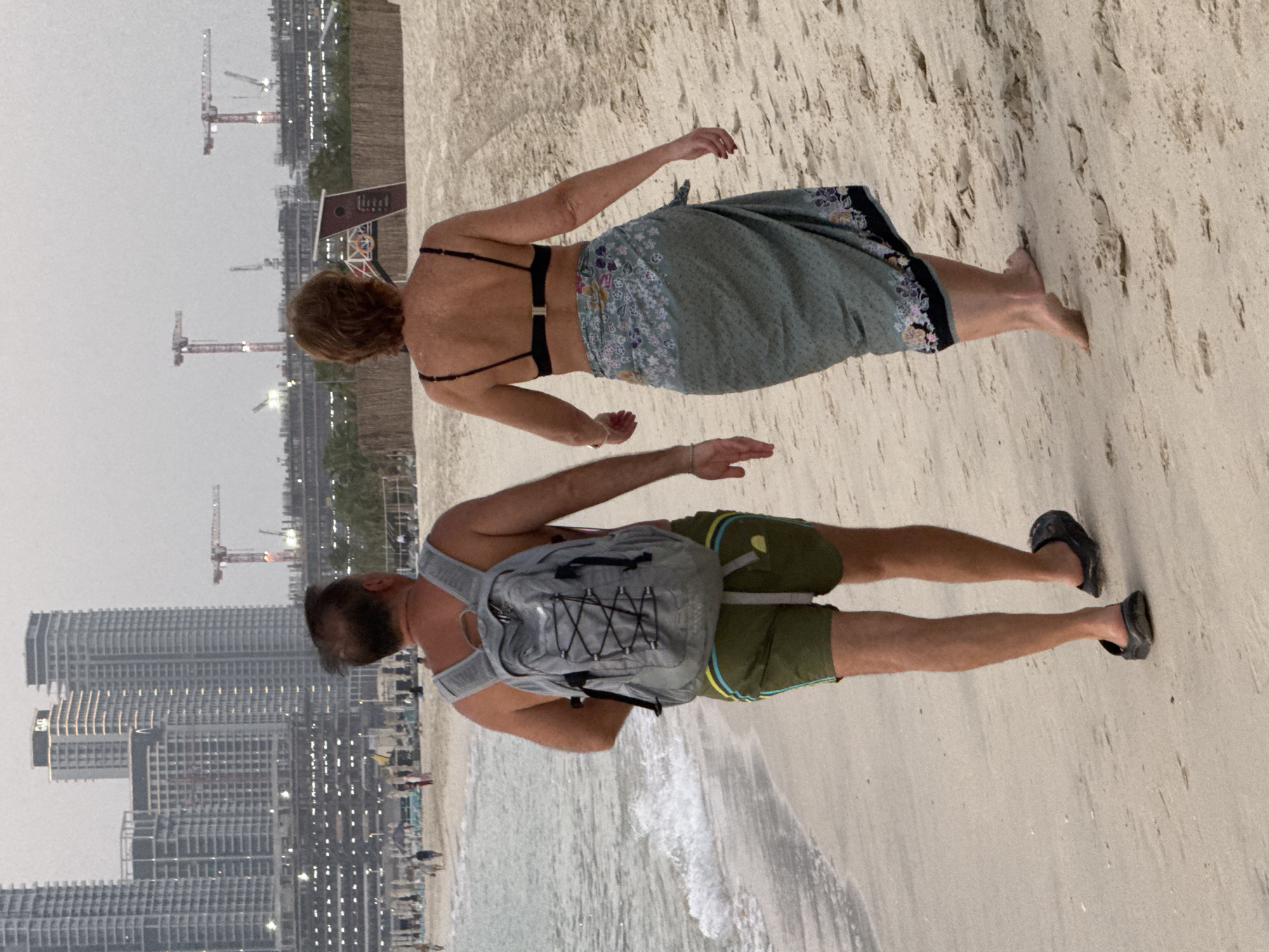 Two people are walking on a beach in Dubai, United Arab Emirates. The man in the foreground is wearing a grey backpack, green shorts, and black sandals. He is walking away from the camera towards the ocean. The woman in the background is wearing a black bikini top and a patterned sarong. She is also walking away from the camera. The beach is sandy, and the ocean is visible to the left. In the background, there are several tall buildings and construction cranes, indicating an urban area under development. The sky is overcast.