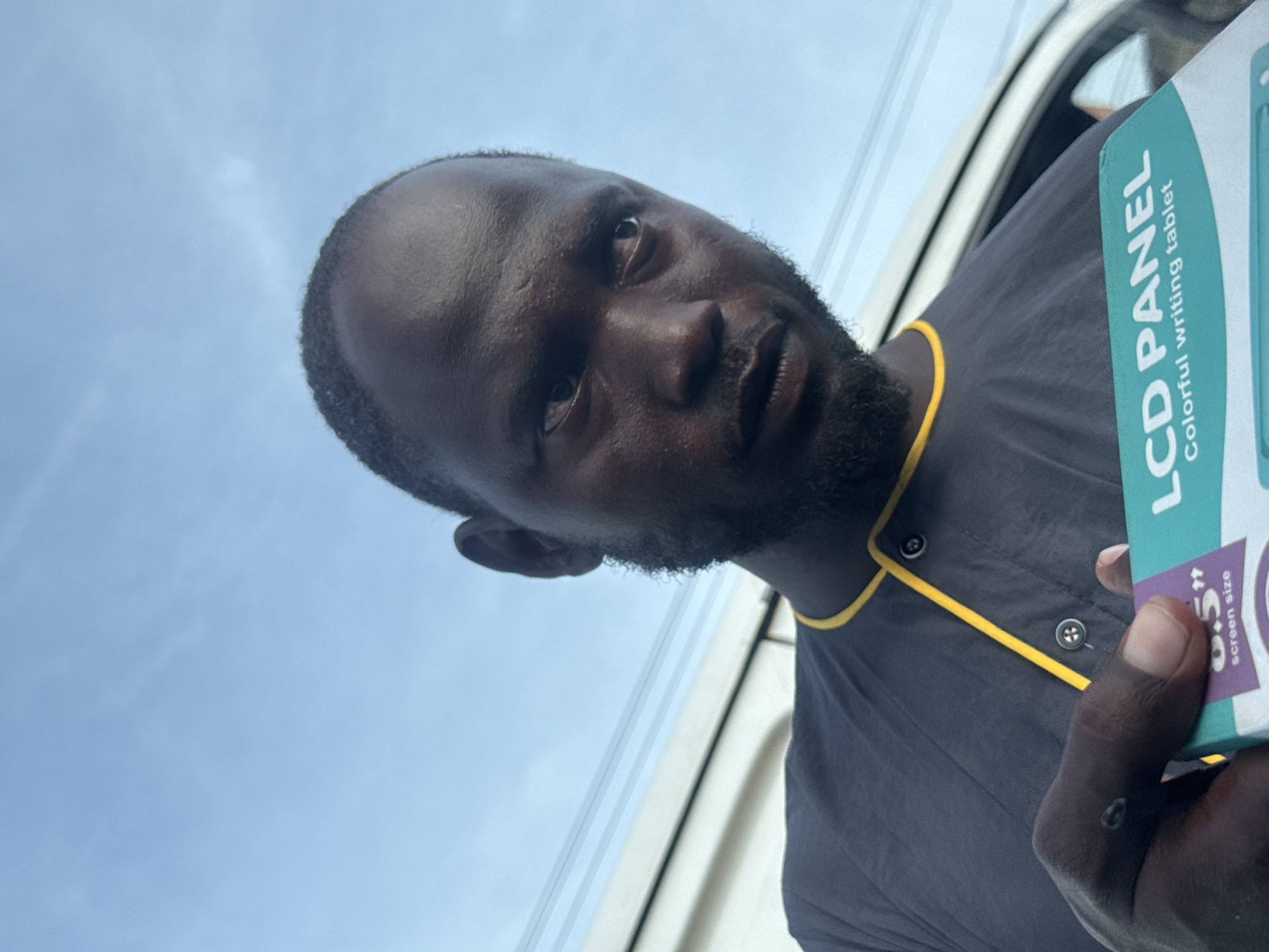 The image shows a dark-skinned man with a beard, holding a box that reads "LCD PANEL Colorful writing tablet". He is wearing a grey shirt with yellow trim. The sky is visible in the background, suggesting an outdoor setting. Based on the prompt, the location is Ado, Nigeria, although no other element in the photo helps confirming this.
