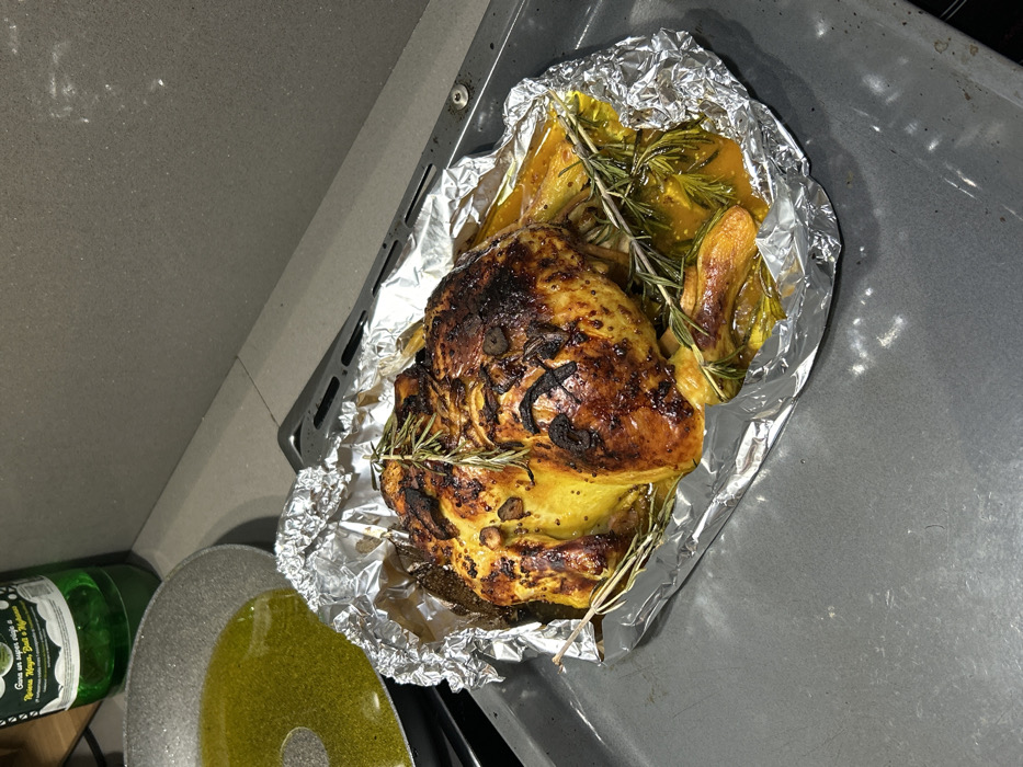 A roasted chicken sits on aluminum foil, accompanied by squash and rosemary sprigs. The chicken's skin exhibits a golden-brown hue, marked with darker patches. Squash pieces are cooked with a glossy appearance. Positioned beside the baking sheet is a green bottle and a pan filled with oil. The setting is a kitchen counter, displaying the ingredients and cooking tools utilized in preparing this dish.