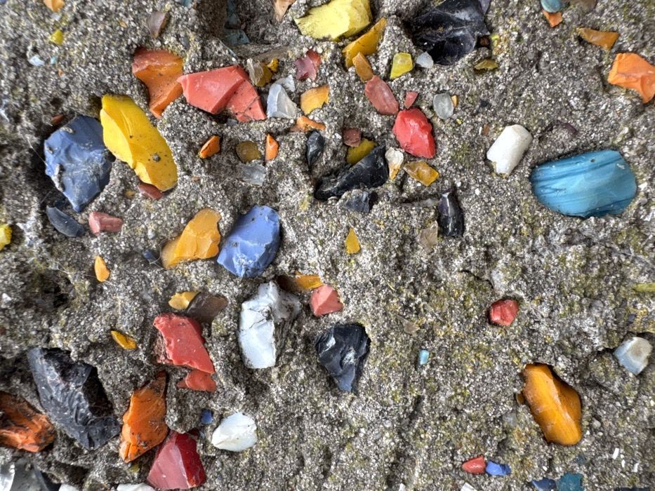 The image displays a close-up, vertical view of a textured, grey, granular concrete-like surface, located in Anapa, Russia. Numerous irregularly shaped fragments are embedded within this surface, varying in size from approximately 1 to 4 centimeters. These fragments consist of opaque and translucent pieces in various colors including red, orange, yellow, blue, black, white, clear, and turquoise, consistent with broken glass or similar vitreous material. The fragments are randomly distributed throughout the surface. The concrete-like material exhibits an uneven, porous texture, with scattered patches of greenish-yellowish discoloration, indicating the presence of moss or lichen. No people, discernible actions, or interactions are visible in the scene.