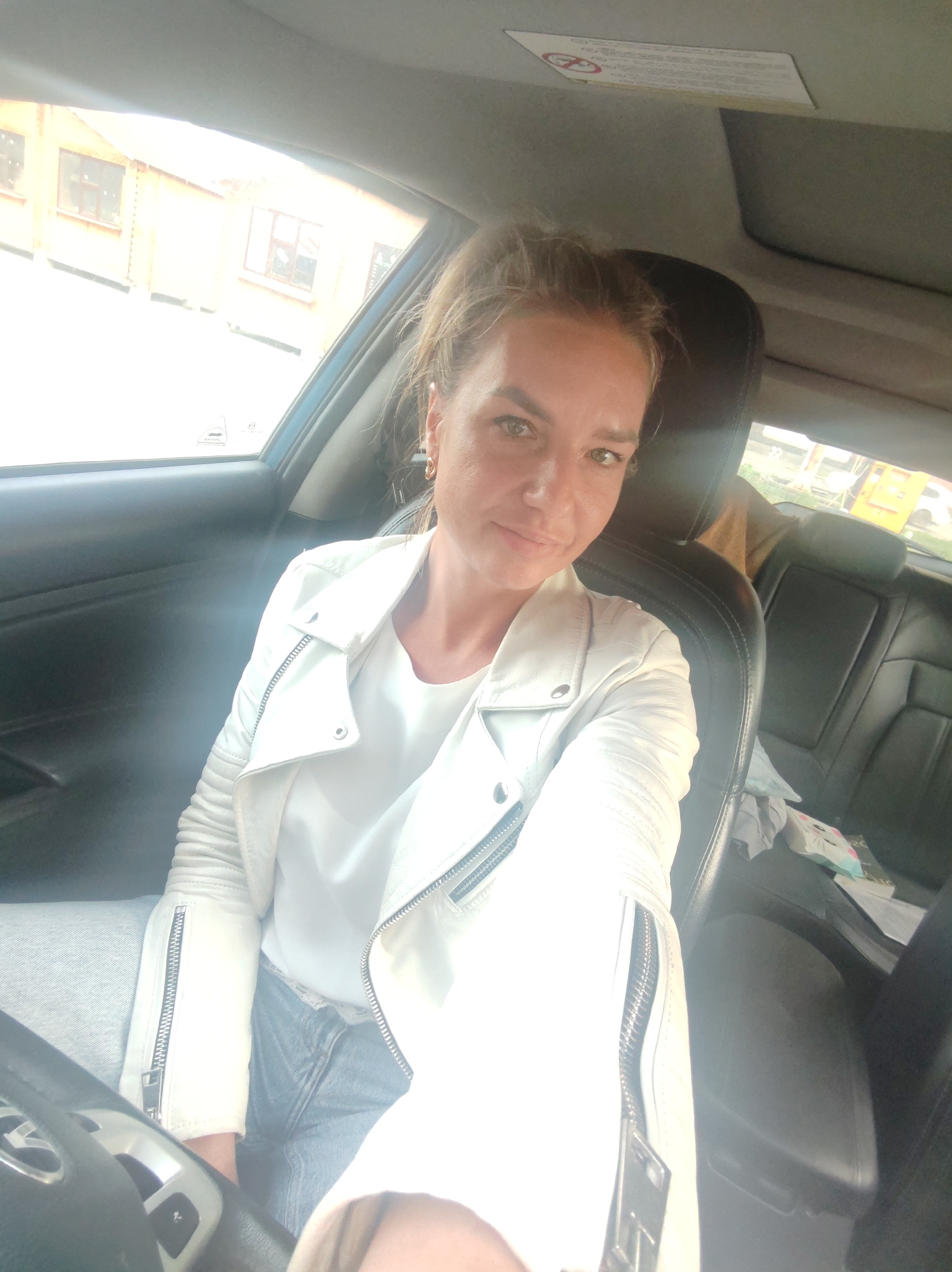 A woman sits in the driver's seat of a car. She is looking at the camera and smiling. She is wearing a white jacket and jeans. The car is parked in front of a building. The lighting in the image is bright, suggesting it is daytime. There are some papers and a small stuffed animal visible in the back seat of the car.