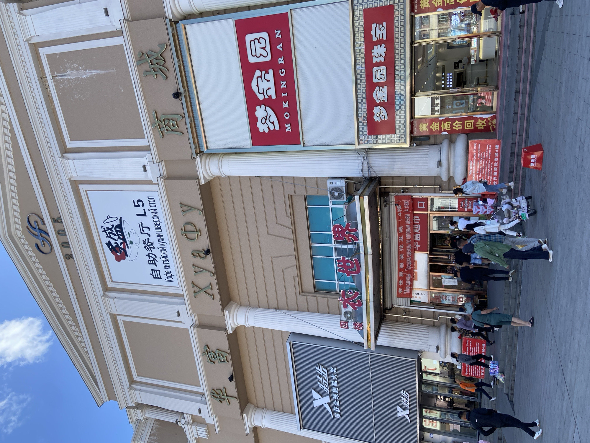 The image captures a street-level view of a commercial building in Xingan Jiedao, China. The building's facade is decorated with ornate architectural elements, including columns and detailed friezes. Several signs are prominently displayed, featuring Chinese characters and some with English text. One large sign on the right side reads "MOKINGRAN" and also displays Chinese characters that translate to "Dream Golden Garden." Another sign above it also appears to be for a jewelry store. A large sign on the left, with the letter "X" and Chinese characters, seems to advertise a brand of clothing or footwear. Below these main signs, storefronts are visible, with one appearing to be a clothing store with racks of garments displayed.
In front of the building, a paved pedestrian area is populated by several individuals. Some are walking, while others are standing or gathered in small groups. One person is seen walking a scooter, and another is sitting on a step. The overall atmosphere suggests a busy commercial district on a sunny day. The sky is visible at the top of the image, showing patches of white clouds against a blue background.
