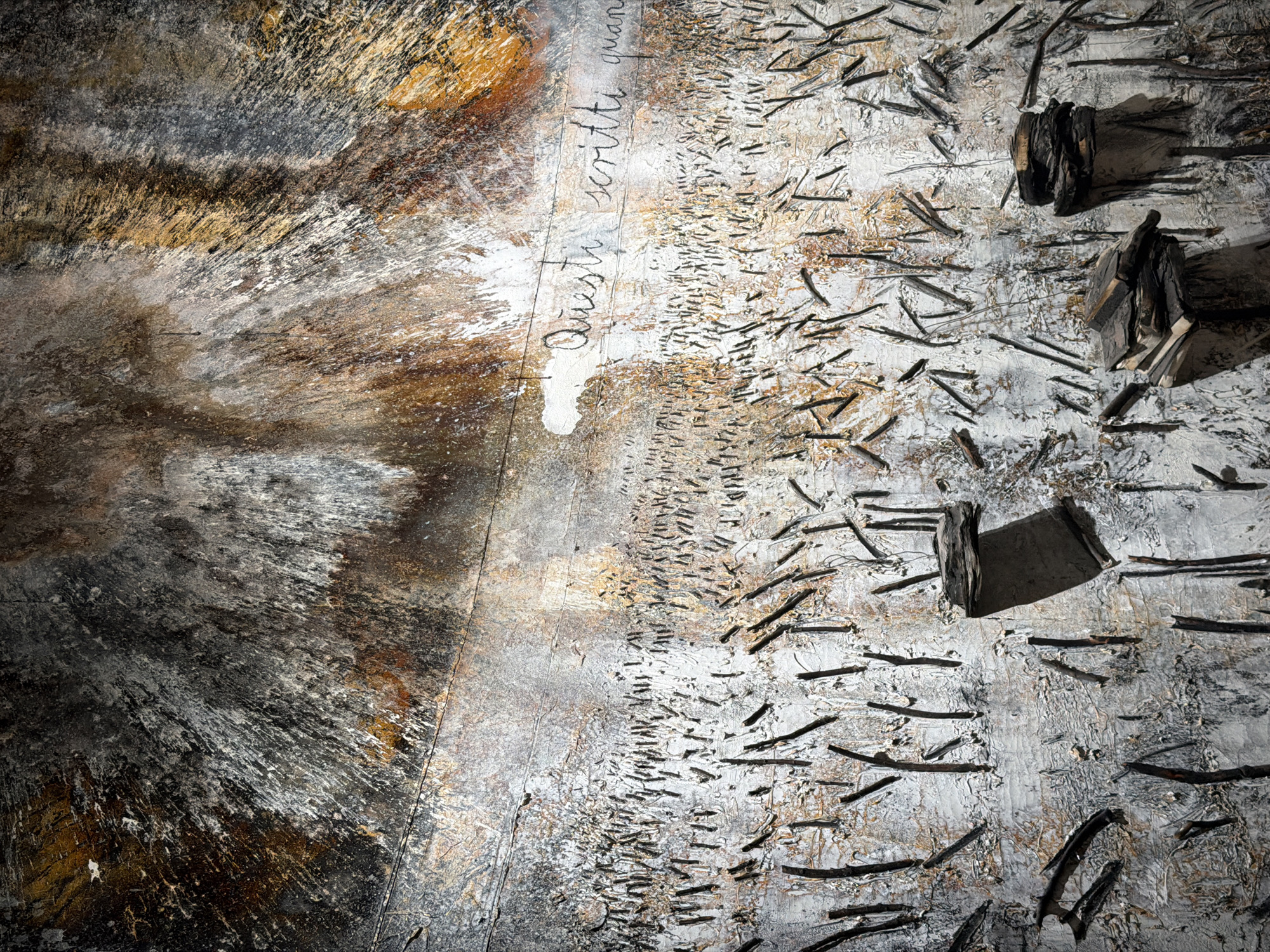 This image displays a textured, abstract artwork with a distressed, layered appearance. The upper portion features a chaotic blend of dark, grey, white, and rusty brown colors, with splatters and streaks suggesting organic or explosive patterns. A horizontal line divides this area from the lower section, which is predominantly textured in white and grey. This lower section is covered in numerous dark, thin, vertical and diagonal slashes or raised elements, resembling dried grass or debris. In the middle ground, faint, handwritten text in black appears on a white patch, reading "Questi scritti quano". In the foreground, there are several dark, three-dimensional elements composed of layered, flat, dark material, resembling small stacks or platforms, some appearing weathered or burnt. The artwork evokes a sense of decay, resilience, or historical marking. The image is a close-up detail of this artwork, and its location in Almaty, Kazakhstan is not identifiable from the visual information provided.