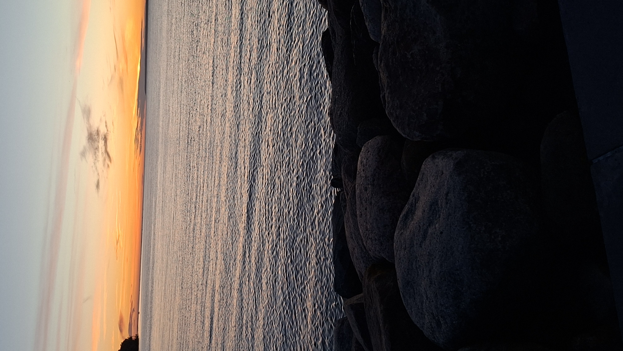 The image depicts a serene waterfront scene at sunset in Tallinn, Estonia. In the foreground, large, dark, and rough-textured boulders line the shore, creating a stark contrast with the water. The water itself is calm, with gentle ripples reflecting the warm hues of the setting sun. The horizon is a brilliant band of orange and pink, characteristic of a sunset, with soft, wispy clouds scattered across the sky. A distant, dark landmass is visible on the left side of the horizon, likely a part of the Tallinn coastline or an adjacent island. The overall atmosphere is tranquil and picturesque, capturing the natural beauty of Tallinn's coastline during twilight.