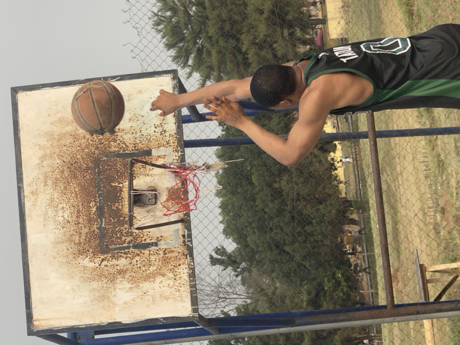 The image displays an outdoor basketball court in Umunekwu, Nigeria. A male individual, viewed from the back and right shoulder, is depicted in the act of shooting a basketball. He wears a black and green sleeveless jersey with "TATUM" and the number "0" visible in white on his back. Both of his arms are extended upwards towards the basketball hoop. A worn, brown basketball is airborne, positioned slightly above and to the right of the hoop's rim. The backboard is a rectangular, white-painted metal structure, extensively covered in brown rust, with the heaviest concentration around the rim's mounting area. The metal rim is rusted, and its attached net, composed of red and white fibers, is tattered and partially torn. The hoop is supported by blue metal posts. A chain-link fence separates the immediate playing area from the background. Beyond this fence, several small, indistinct human figures are visible in an open, grassy expanse. Numerous green trees form the backdrop, and a single bare tree branch is noticeable on the far left. A rudimentary wooden structure, possibly a bench or table, is partially visible in the lower-left foreground, behind the chain-link fence. The sky above is overcast.