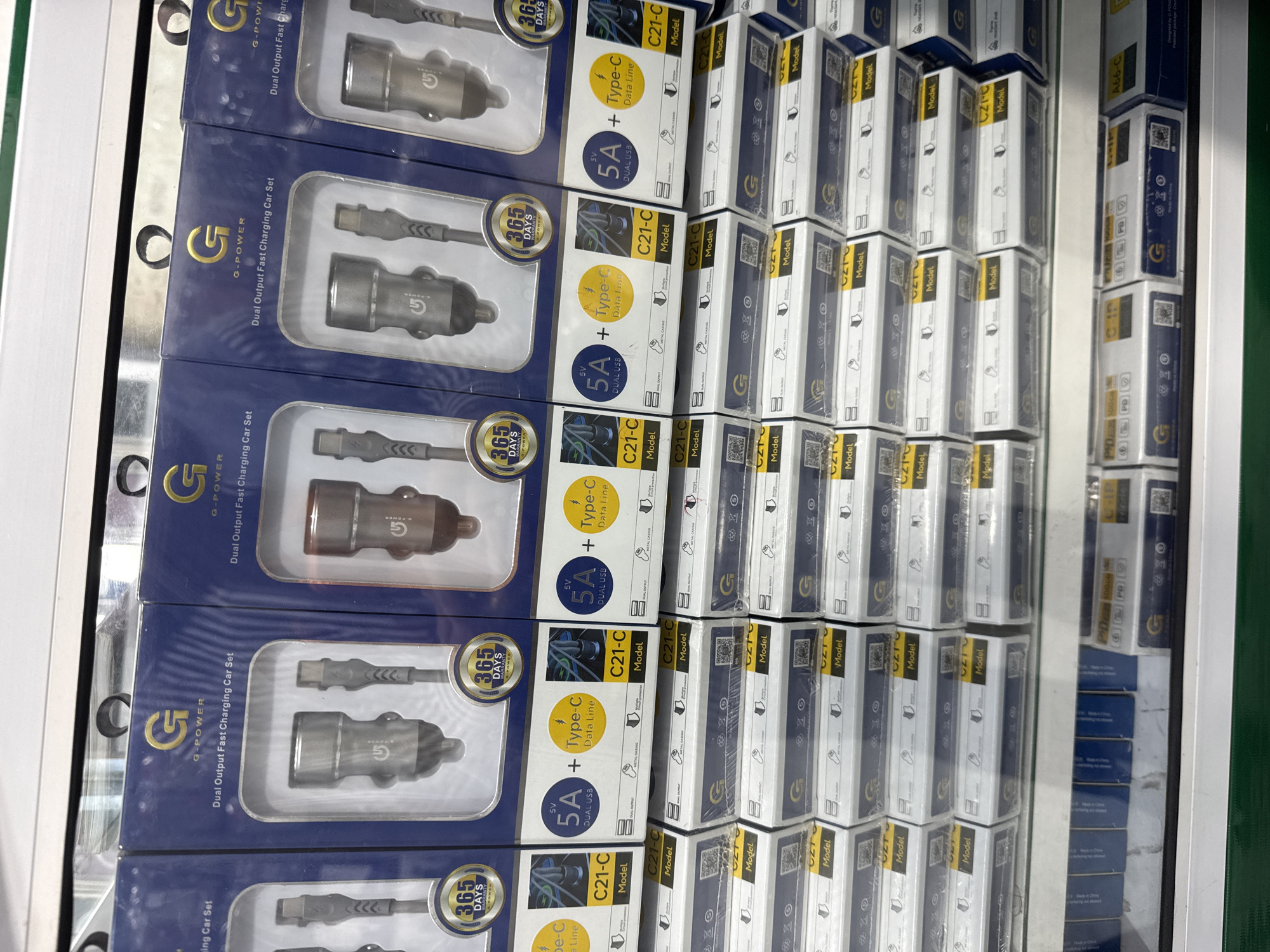 The image shows a retail display of mobile phone accessories, specifically car chargers and data cables. The products are neatly arranged in stacks, predominantly in their original packaging. The packaging is primarily dark blue with gold and white accents, featuring branding that includes a prominent "G" logo and the text "G-POWER" and "Dual Output Fast Charging Car Set". Some packages also highlight "365 DAYS WARRANTY" and specifications like "5V 5A DUAL USB" and "Type-C Data Line". The model numbers "C21-C" and "A66-C" are also visible. The background suggests a store or warehouse setting with shelves and fluorescent lighting, typical of a point-of-sale environment. There are no people visible in the image, and the overall impression is one of commercial organization and product presentation. The visible text on the packaging indicates the product type, brand, warranty information, and technical specifications. The location context of Bireuenmeunasah, Indonesia, is provided as external information.