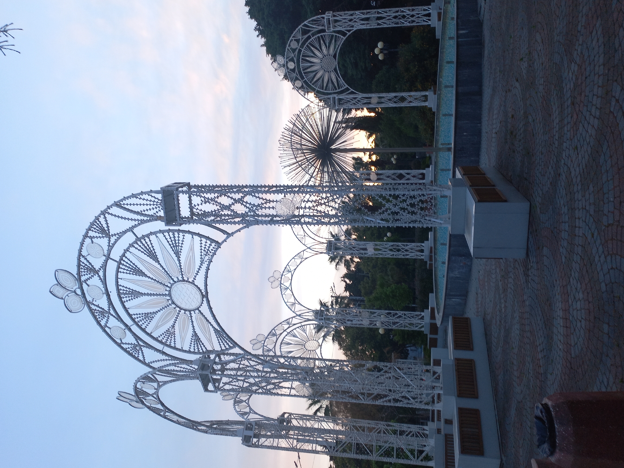 The image depicts a series of decorative, illuminated archways in what appears to be a park or public space, possibly in Sochi, Russia. The archways are constructed from a light-colored, possibly white, metal framework with floral and geometric designs. Behind the archways, there is a backdrop of green foliage and trees, with a glimpse of the sky visible through the structures. The lighting suggests it is either early morning or late evening. The ground is paved with a patterned brick or stone. There are benches along the side of the archways. There are no people visible in the frame.