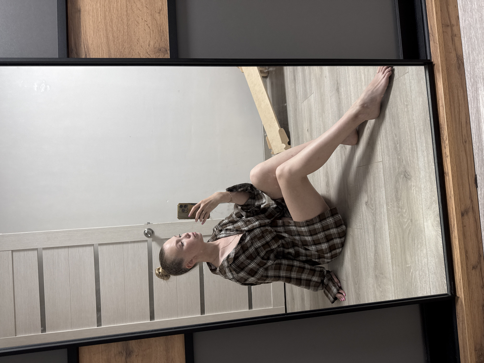 A young woman with blonde hair in a bun is sitting on a wooden floor in a room, taking a selfie in a mirror. She is wearing a brown and white plaid oversized shirt. Her left hand is raised, holding a phone. Her legs are extended in front of her. The room appears to be a bedroom or dressing area, with a door visible on the left and a wooden textured wall section on the right. There is a light-colored wooden ramp-like object in the reflection of the mirror. The floor is made of light-colored wood planks. The overall lighting is neutral. The image is taken from a slightly elevated angle.