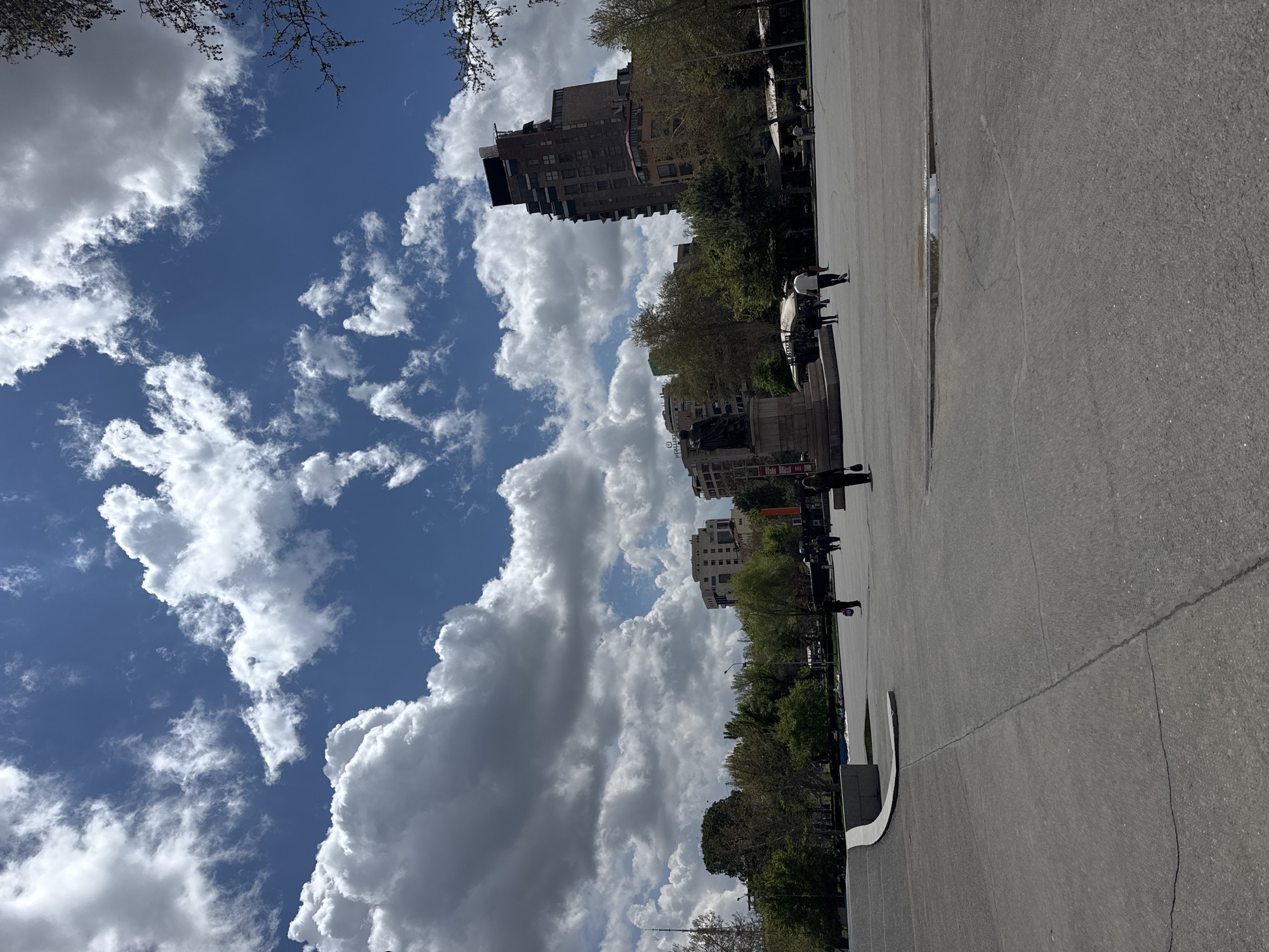 This image depicts a wide, brightly lit urban scene, likely a public square or park in Yerevan, Armenia. The photograph is taken on a sunny day, characterized by a vibrant blue sky dotted with numerous fluffy white cumulus clouds.
The foreground and much of the midground are dominated by a vast expanse of grey paved ground, resembling asphalt or concrete. Near the center-left of this paved area, a reflective puddle is visible, suggesting the presence of standing water. Towards the bottom-left, a low, white curved barrier or structure marks the edge of the paved surface.
In the background, a row of trees with emerging green foliage separates the open ground from several multi-story buildings. These buildings display varying architectural styles, some featuring balconies. Amongst the trees and buildings, a dark-colored, prominent statue or monument stands on a discernible base, hinting at the area's cultural or historical significance. Distant figures of people are scattered across the paved area and near the background elements, appearing to be walking or standing, though their specific activities are indistinct due to distance.
The bright sunlight suggests it is midday or early afternoon. Tree branches with sparse leaves frame the upper edges of the sky. Text, possibly in Armenian script, is visible on the base of the monument, but it is too blurry to be deciphered.