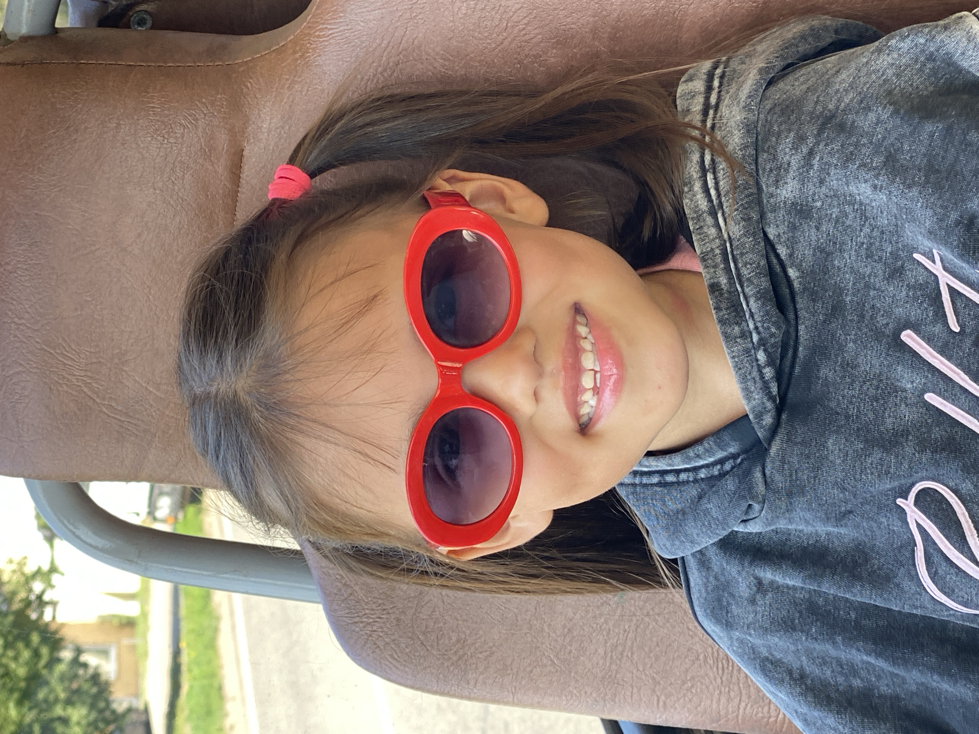 A young girl with dark hair tied in pigtails with pink elastics, wearing red oval sunglasses and a dark grey hoodie, sits in a brown upholstered seat, possibly in a vehicle. Her mouth is open in a wide smile, revealing her teeth. The background is blurred, showing a hint of green foliage and what appears to be a building, suggesting an outdoor setting in a residential area of Chaikovsky, Russia. The lighting indicates it is daytime.