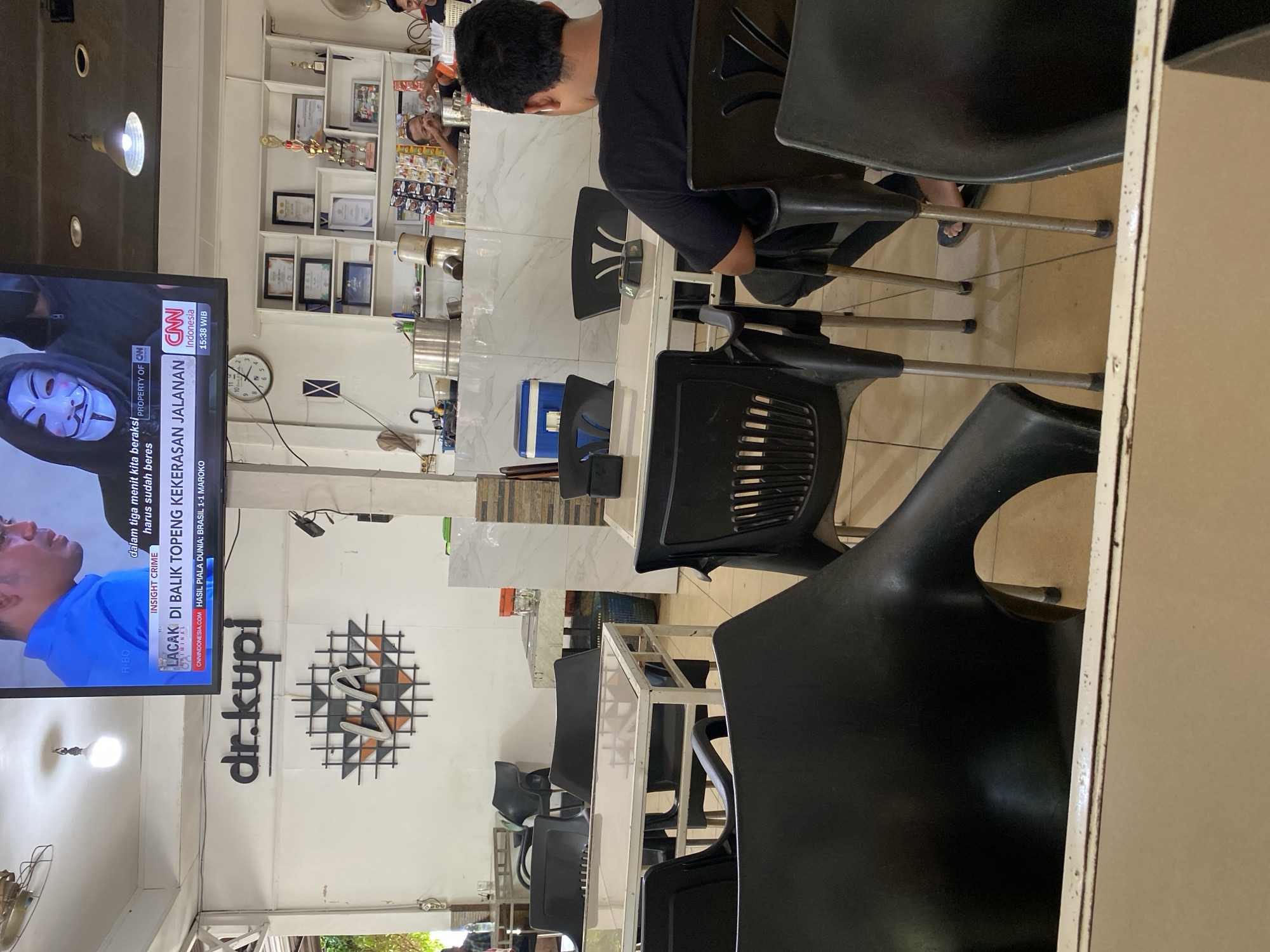 The image depicts the interior of a cafe, likely "dr.kupi," judging by the prominent signage on the wall. The cafe appears to be in Lhokseumawe, Indonesia, as suggested by the CNN Indonesia broadcast visible on the television screen, which displays news headlines in Indonesian. The cafe has a casual, indoor seating arrangement with multiple tables and black plastic chairs.
In the foreground, a person wearing a black t-shirt and dark pants is seated facing away from the camera, looking towards a table. Their feet, clad in dark flip-flops, are visible resting on the tiled floor. Behind this person, another black plastic chair is partially visible.
The cafe's environment includes white walls, some decorated with a geometric wall art piece. Shelving units are present in the background, displaying framed certificates, trophies, and various items, suggesting the cafe may also have awards or recognition displayed. A television screen mounted on the wall is showing a news broadcast from CNN Indonesia, with Indonesian text and imagery of people in masks and a news anchor.
On a counter area in the mid-ground, various items are visible, including stainless steel containers, what appears to be a blue cooler, and stacked items that might be food or drinks. Two individuals are partially visible behind this counter, one of whom is interacting with items on the counter. The overall lighting suggests an indoor setting, possibly during the daytime. The flooring is tiled.