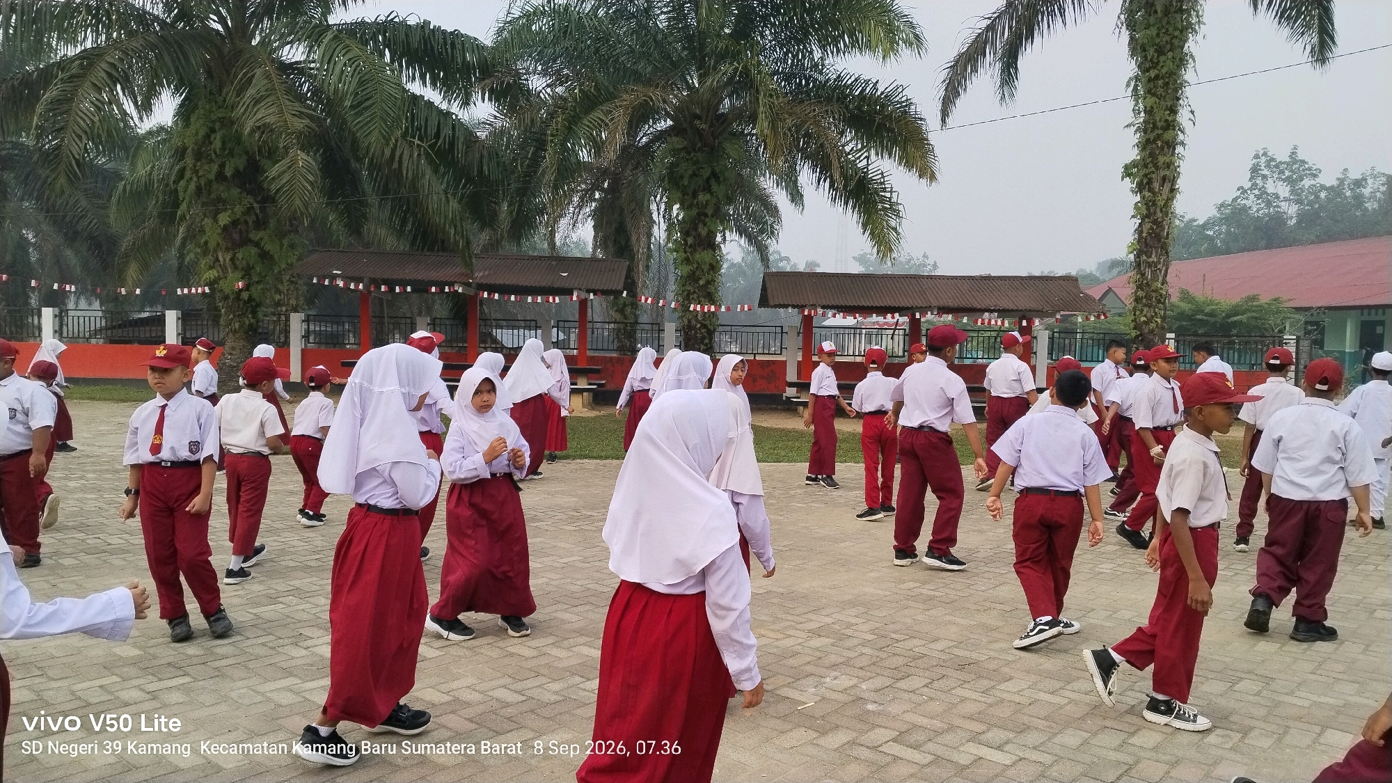 In an outdoor setting at "SD Negeri 39 Kamang, Kecamatan Kamang Baru, Sumatera Barat, Indonesia," a group of elementary school students, both boys and girls, are engaged in an activity on a paved courtyard. The students are dressed in school uniforms consisting of white shirts, red pants or skirts, and some are wearing red caps. Several girls are also wearing white headscarves. The students appear to be in motion, with some walking and others standing. In the background, there are palm trees, a building with a red roof, and covered seating areas decorated with red and white flags. The overall atmosphere suggests a school event or physical activity taking place outdoors. The image is dated September 8, 2026, at 07:36.