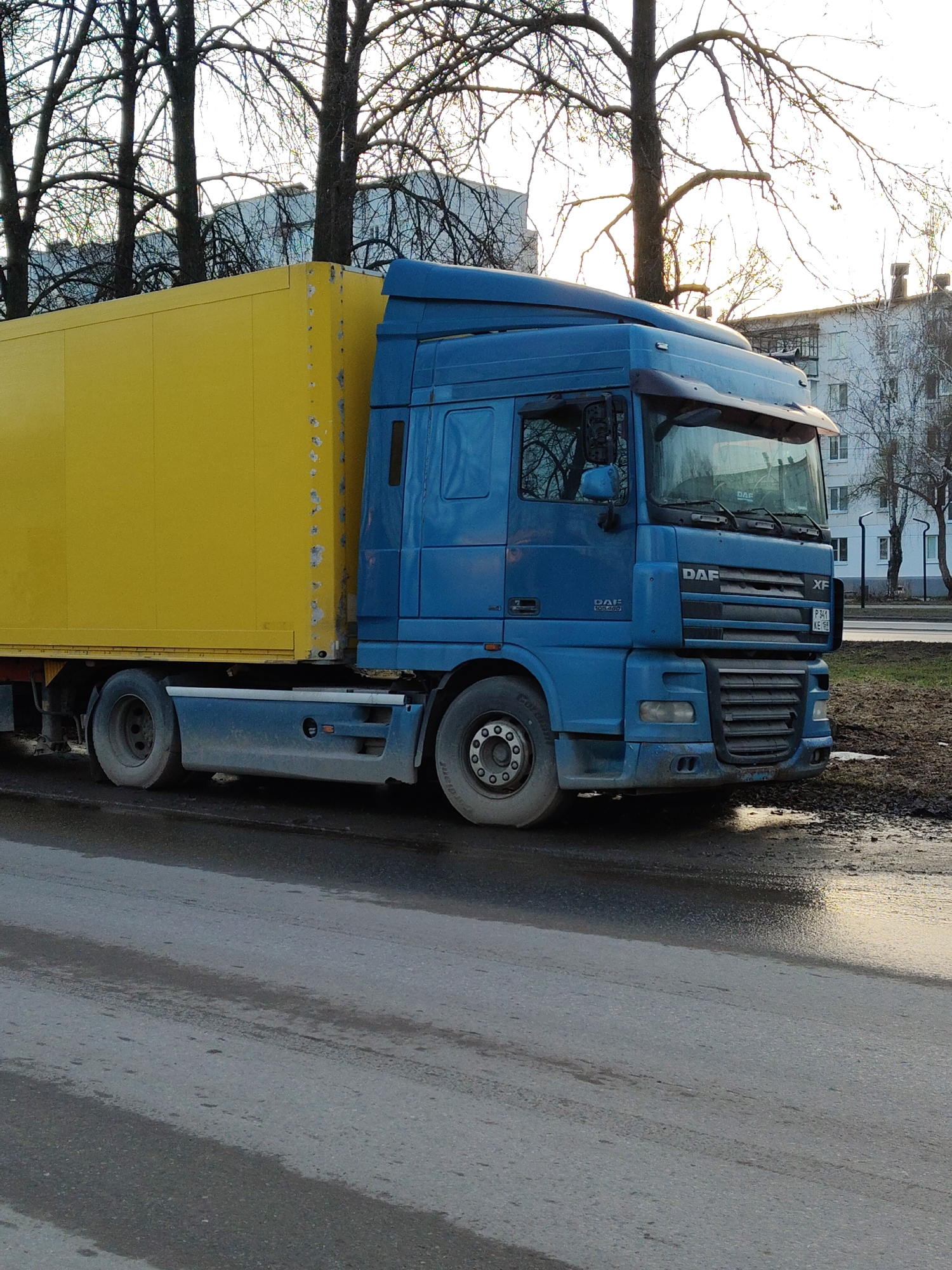 A blue DAF XF semi-truck with a yellow cargo trailer is parked on a city street. The truck is angled slightly towards the viewer, revealing the front right side of the truck. The truck has a license plate that reads "P 341 KE 65". The trailer is positioned behind the cab, and the road is wet with puddles, indicating recent or ongoing precipitation. Bare trees are visible in the background, along with buildings in the distance. The location appears to be an urban area. The city is unknown.