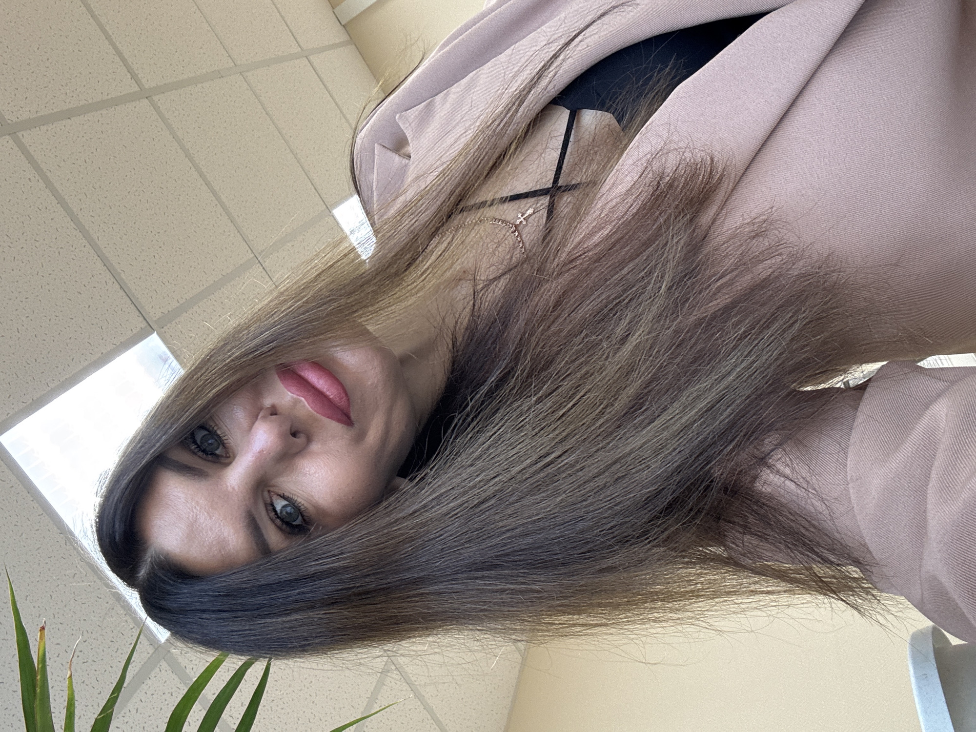 An over-the-shoulder, selfie-style portrait captures a woman with long, dark brown hair and striking blue eyes. She is wearing a light pink blazer over a black top and a delicate gold cross necklace. Her lips are painted a vibrant pink. The background suggests an indoor setting, possibly an office, with a white tiled ceiling and fluorescent lighting fixtures visible. A portion of a green plant with long leaves enters the frame from the upper left corner. The lighting is bright, indicating daytime. The overall impression is casual yet polished. The image was taken in Chaikovsky, Russia.