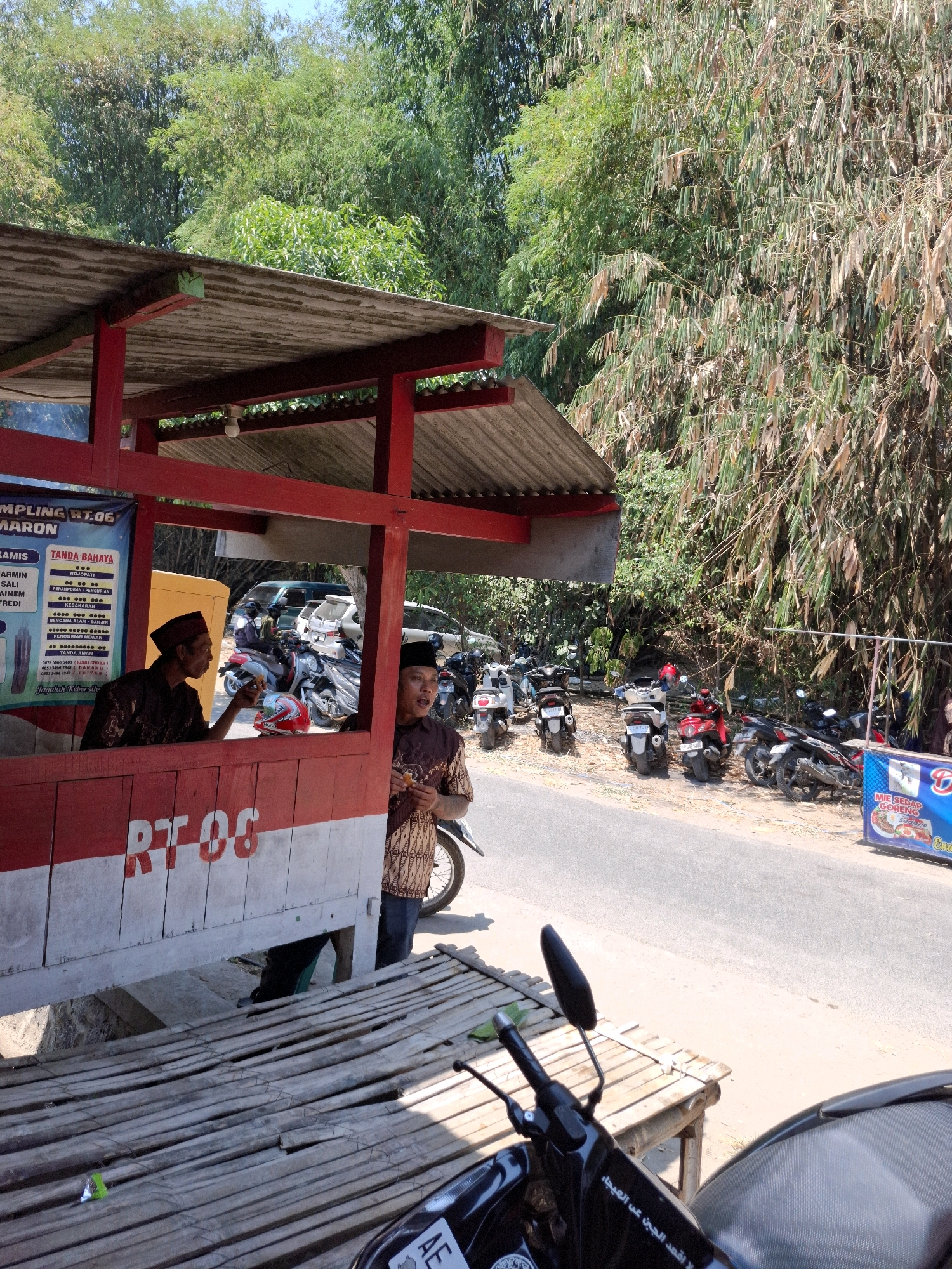 The image shows a street scene in Kedungmaron, Indonesia, with a red and white roadside stall in the foreground. Two men, dressed in traditional Indonesian attire, are interacting at the stall. The man on the left is wearing a dark red patterned shirt and a peci hat, and appears to be speaking to the man on the right. The man on the right is wearing a brown and white patterned shirt and a peci hat, and is holding a piece of food. Behind the stall, a row of parked motorcycles is visible, along with a road. The background is dominated by lush green bamboo trees, suggesting a natural or semi-rural setting. The stall has a corrugated metal roof and wooden construction, with text and graphics on its side. A sign on the stall reads "RT 06". The overall atmosphere is bright and sunny, indicating daylight.